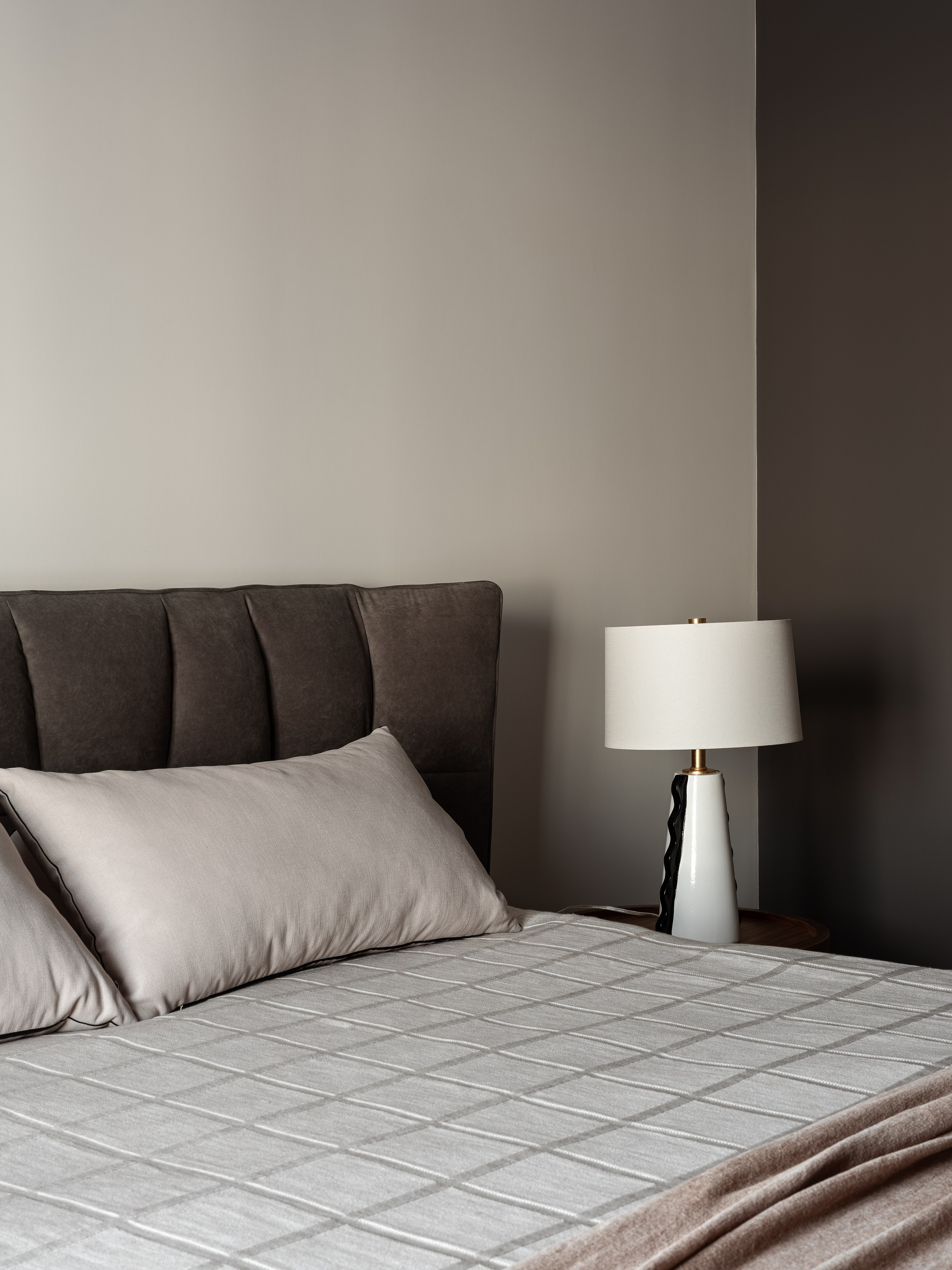 Уютная спальня с серым текстильным изголовьем и декоративными подушками, прикроватная лампа на деревянной тумбе, дизайнер Екатерина Мищенко, фотограф Сергей Денисенко, современный интерьер в пастельных тонах, вид сбоку на кровать.