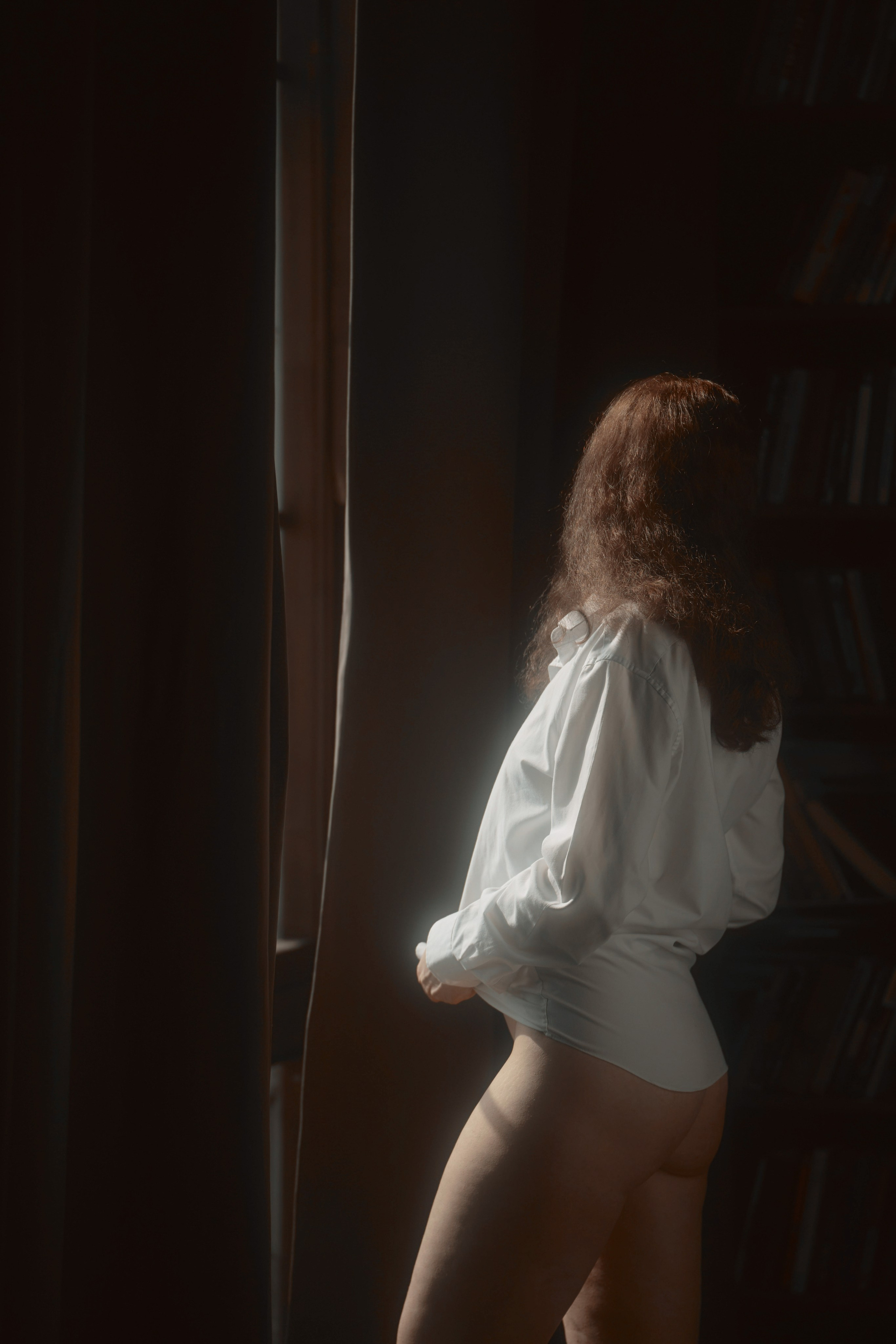 девушка в мужской рубашке стоит у окна напротив стелажа с книгами