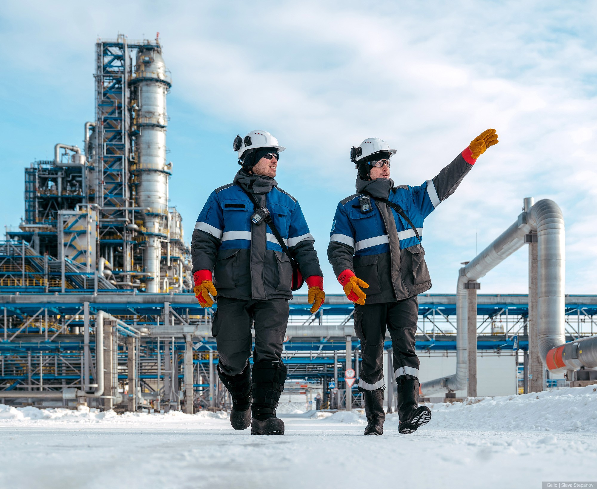 Два инженера в зимней спецодежде обходят территорию газоперерабатывающего завода, специалисты в касках на фоне огромных ректификационных колонн и заснеженных эстакад, Gelio.