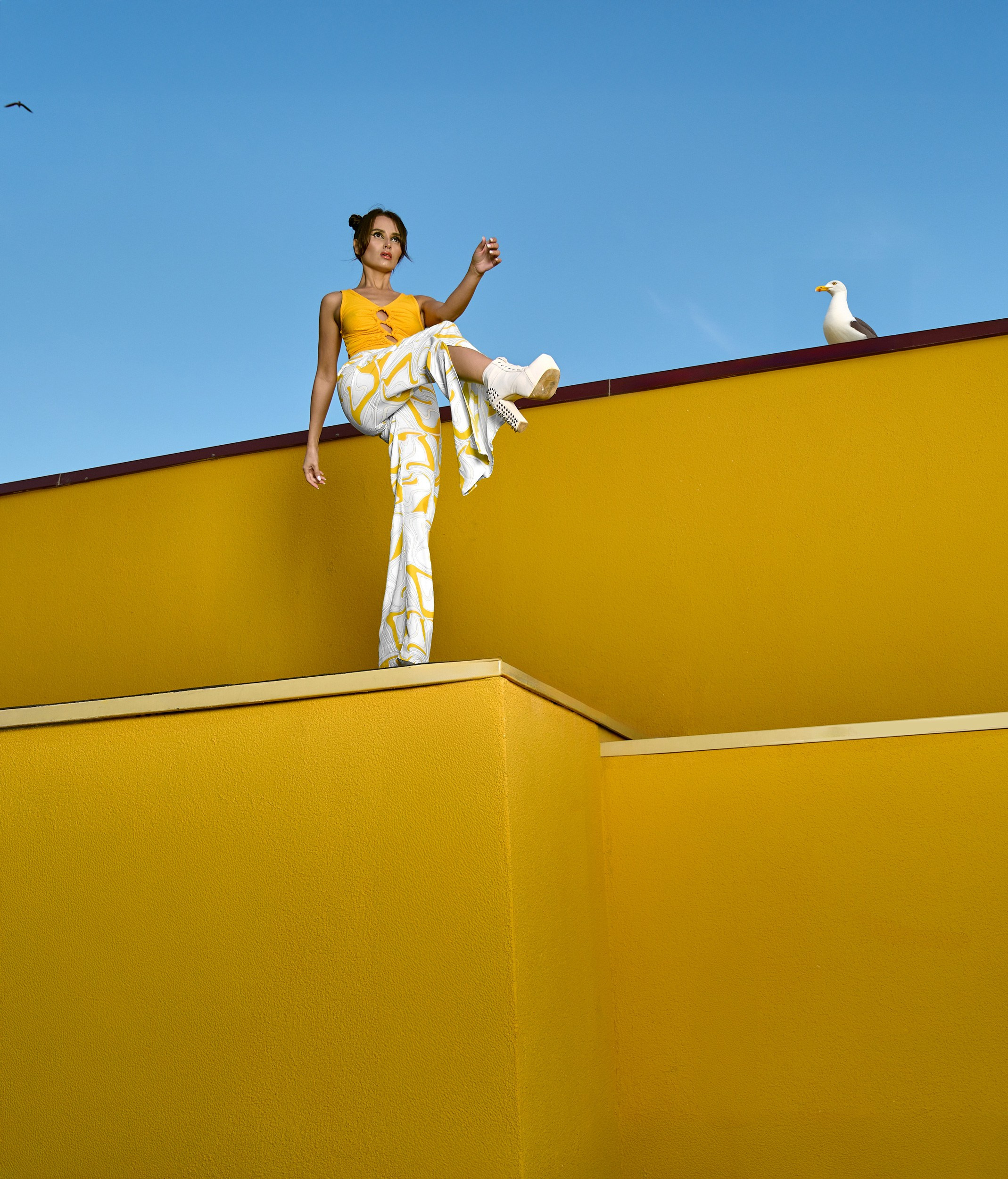 Модель сидит на краю желтой стены в оранжевом топе и белых брюках с желтым принтом. Морская чайка на крыше добавляет элемент естественной композиции
