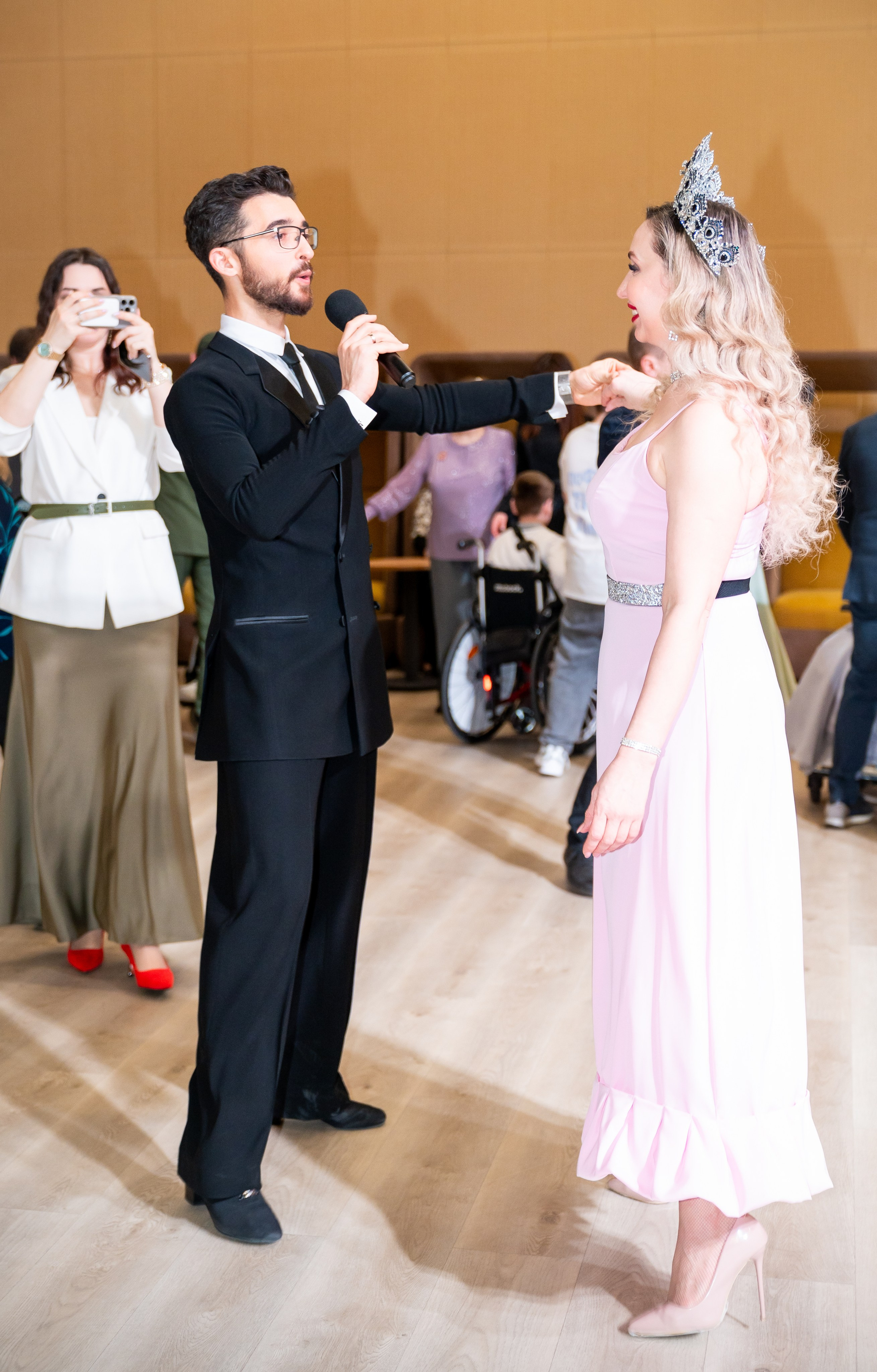 Ведущий в чёрном костюме с микрофоном представляет девушку в розовом платье и короне на инклюзивном балу проекта «Мы все равны». Фотограф Мальцев Евгений.