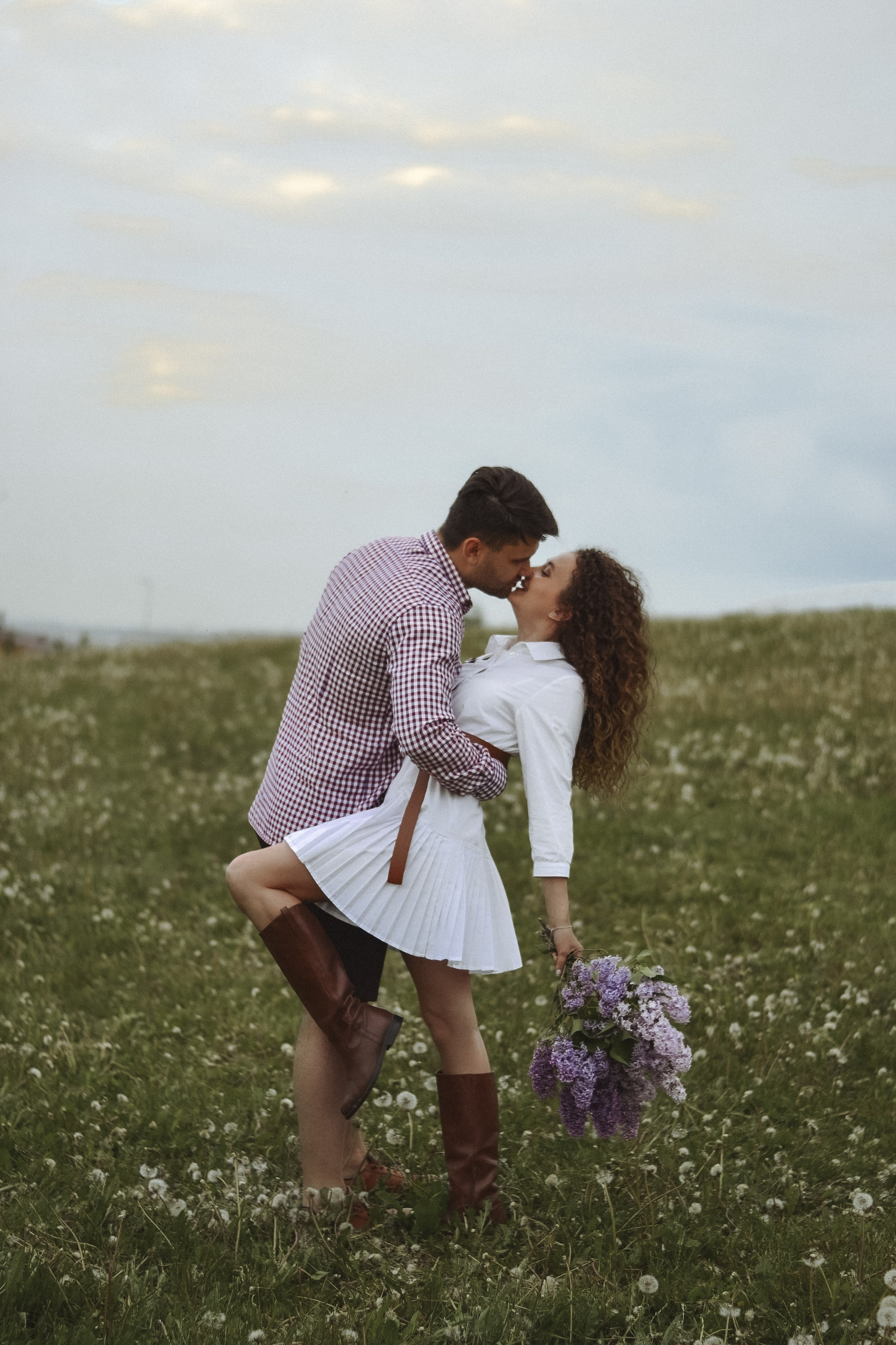 На фотографии изображена пара, обнимающаяся на фоне поля с цветущими растениями. Мужчина одет в клетчатую рубашку и темные штаны, а женщина - в белую юбку и коричневые сапоги. У нее в руках букет фиолетовых цветов. Они стоят лицом друг к другу, и кажется, что они целуются. Это романтическое изображение, которое может быть частью свадебной фотосессии или просто моментом близости между двумя людьми.