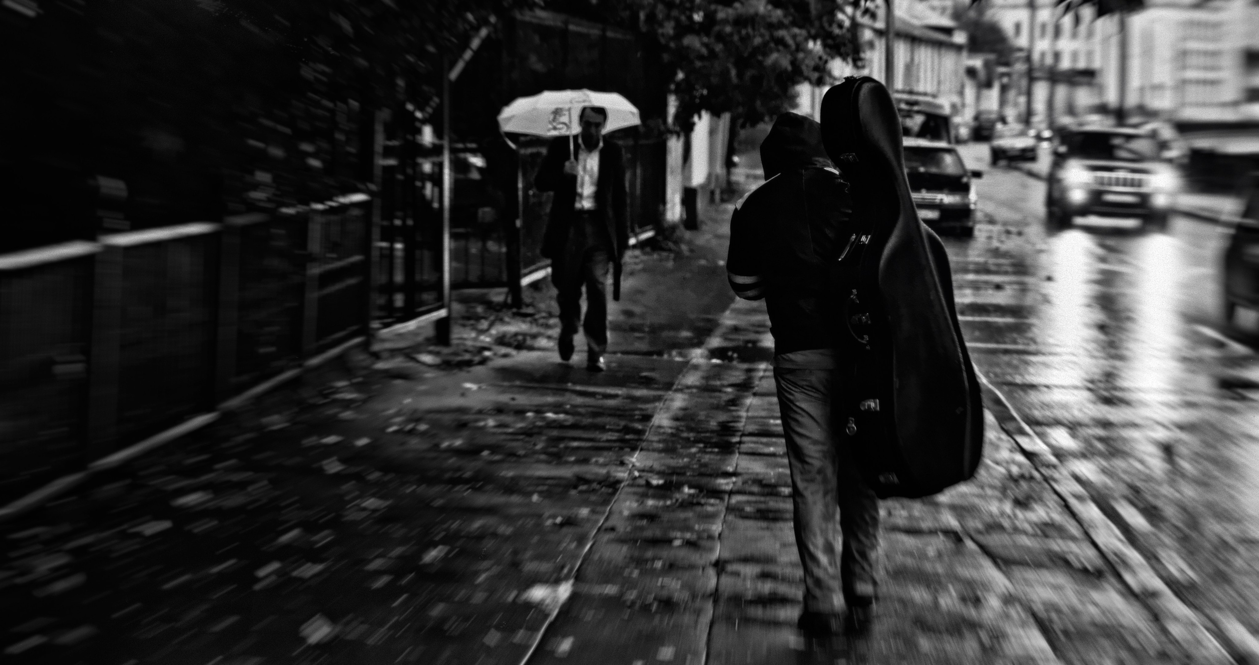 Человек с чехлом для гитары идущий по дождливой улице, размытие движения, человек с зонтом на фоне, атмосферное уличное фото