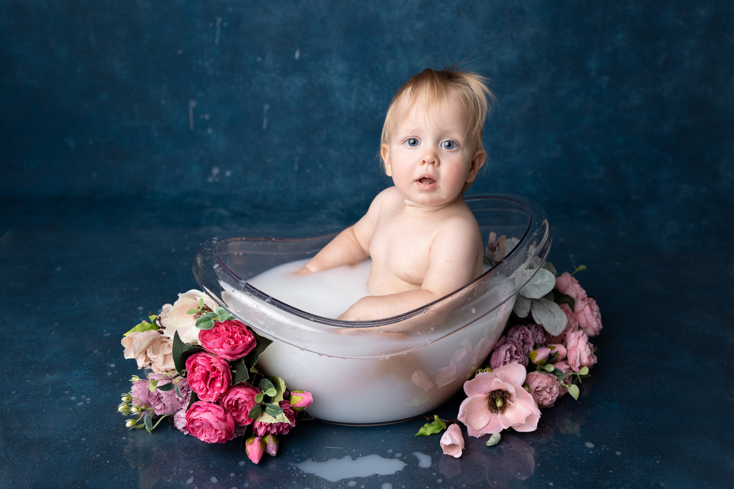 Младенец в прозрачной ванночке с молоком, окруженный цветами, на синем фоне.