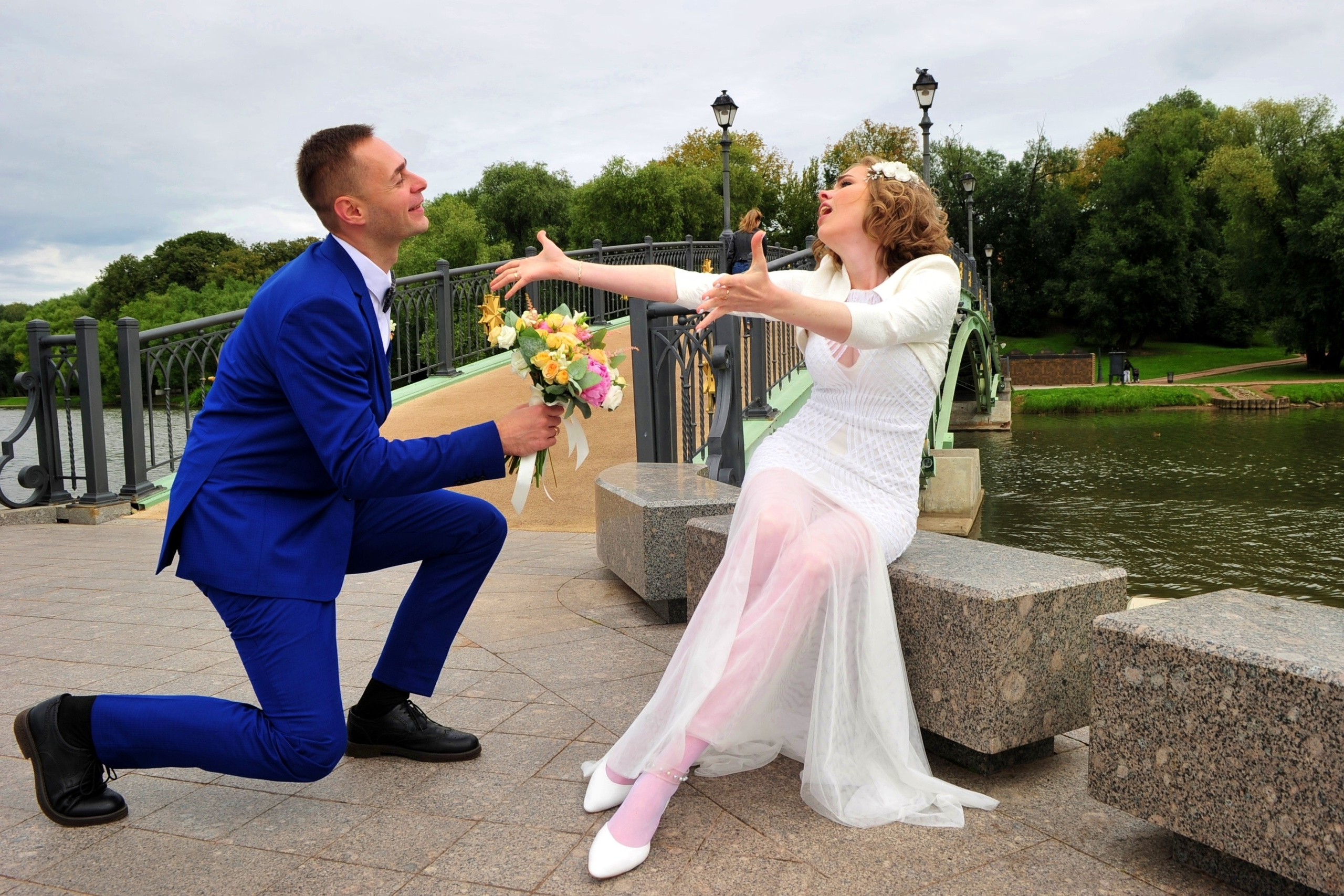 Свадьба в Царицыно хмурым прохладным летним днём, жених стоит на одном колене перед невестой и дарит ей букет, невеста протягивает руки навстречу жениху, новобрачные на фоне пруда у моста