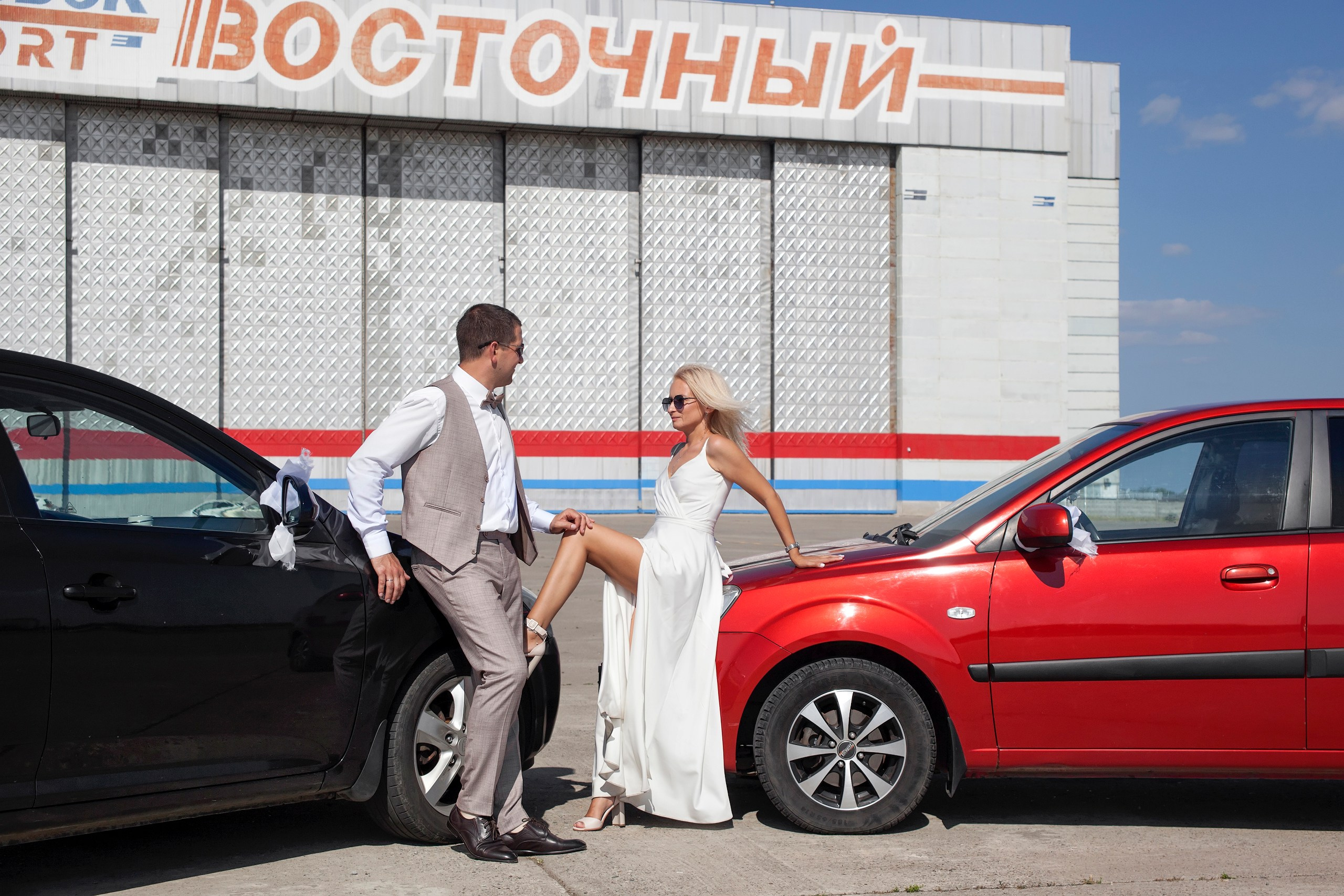 Жених сидит на капоте черной машины, а невеста на капоте красной и опирается ногой на колесо черной машины