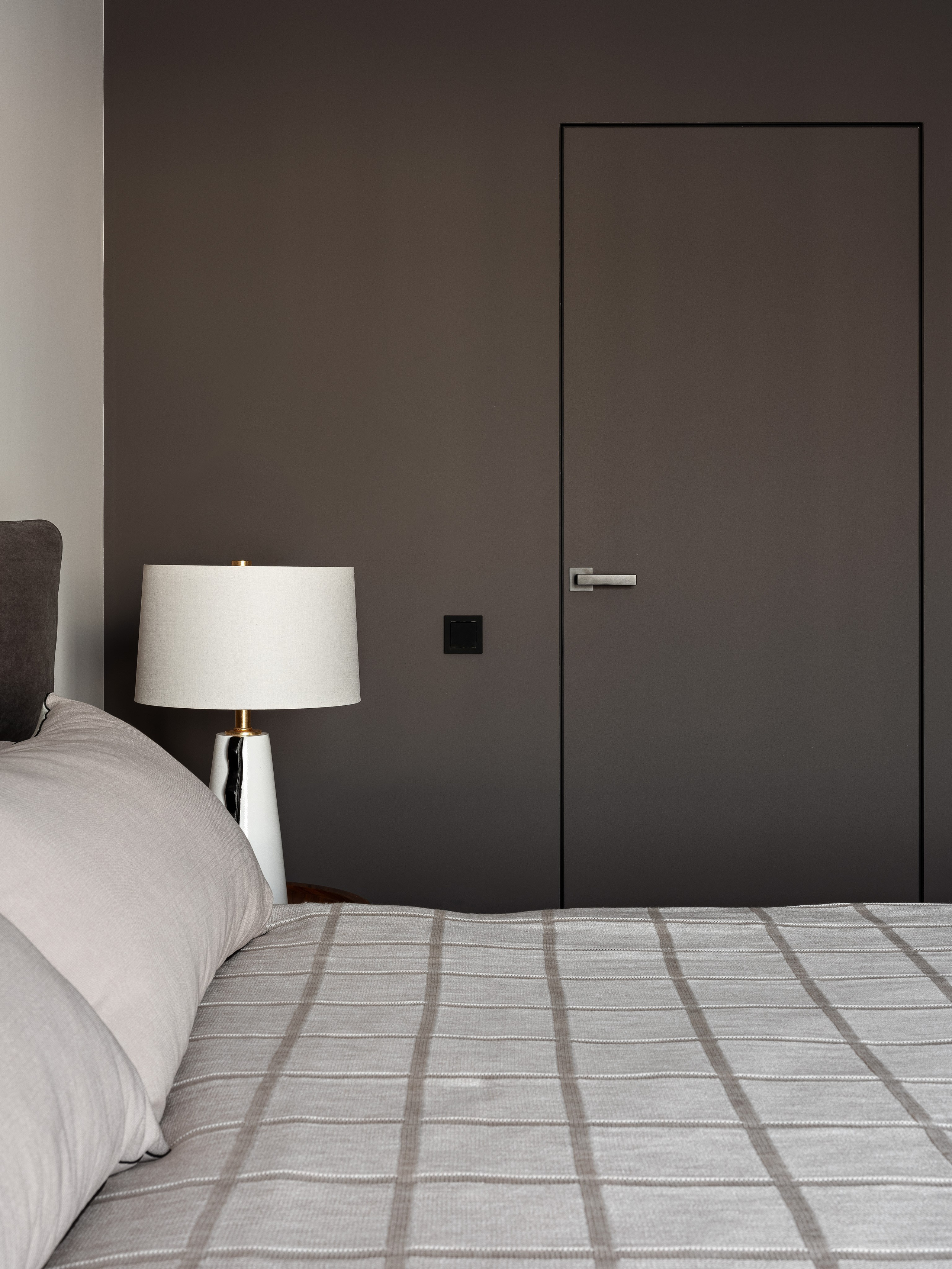 Стильная спальня с серой стеной и скрытой дверью, крупная кровать с мягким изголовьем, прикроватный светильник на деревянной тумбе, дизайнер Екатерина Мищенко, фотограф Сергей Денисенко, современный интерьер в нейтральных оттенках.