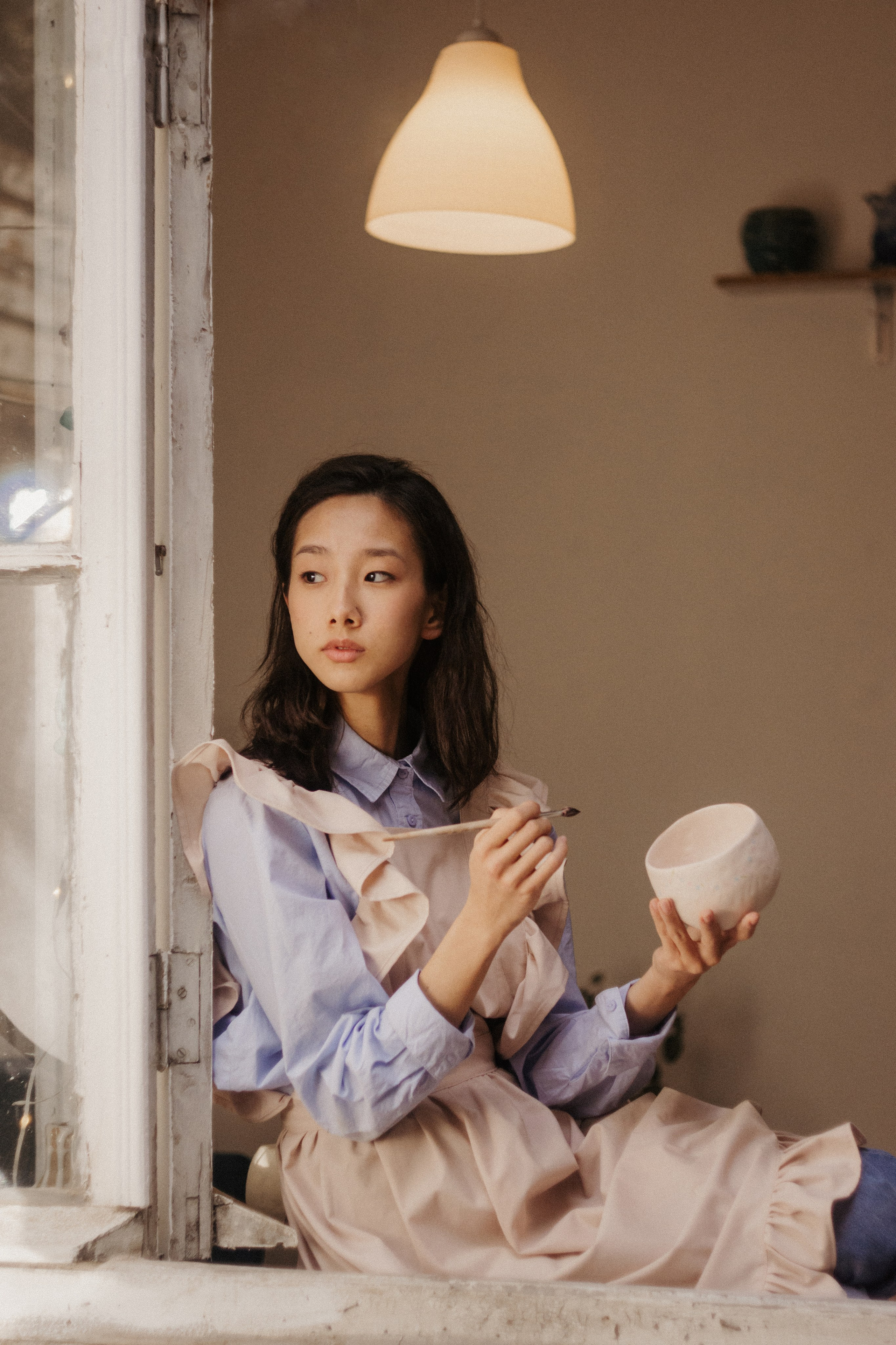 Девушка сидит на подоконнике и держит в руках кисть и глиняную тарелку. У нее темные волосы, светло-голубая рубашка и объемный светло-розовый передник