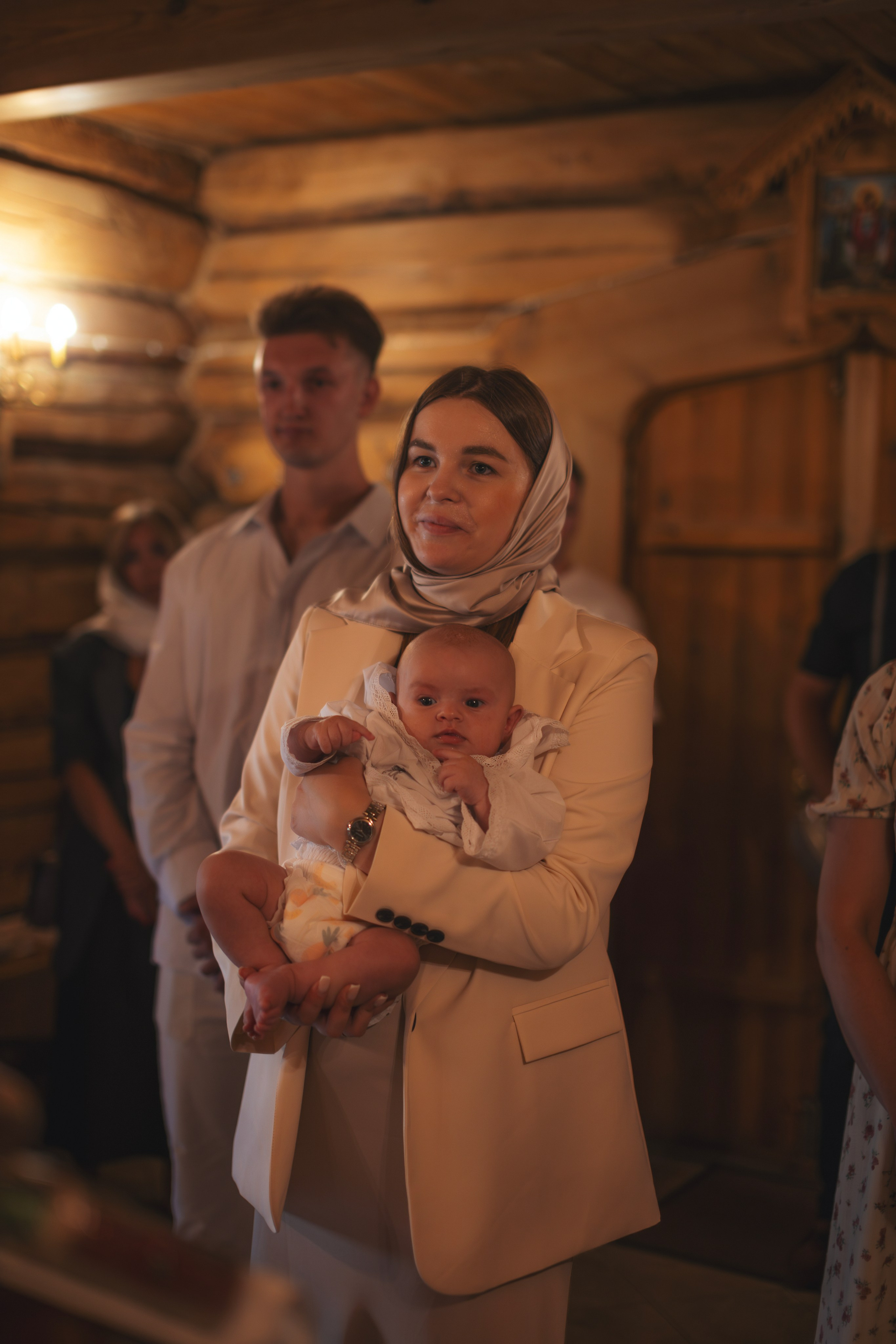 Мама держит младенца во время крещения в церкви Успения Пресвятой Богородицы, окружающие наблюдают за обрядом. Женщина в светлом костюме и платке, ребенок в крестильной одежде.