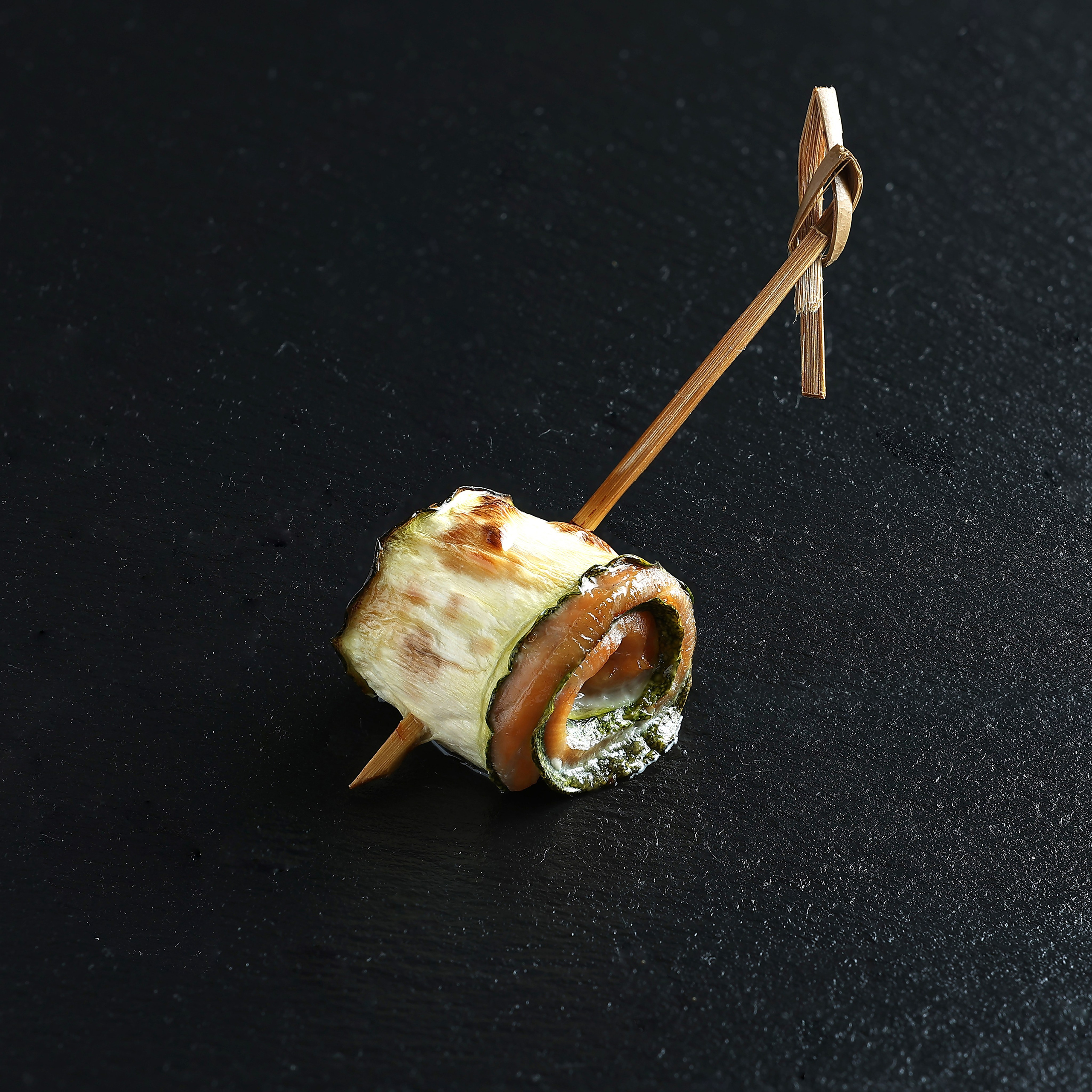 Закуска-рулет на шпажке из обжаренного цукини и мяса с соусом, на тёмном фоне.