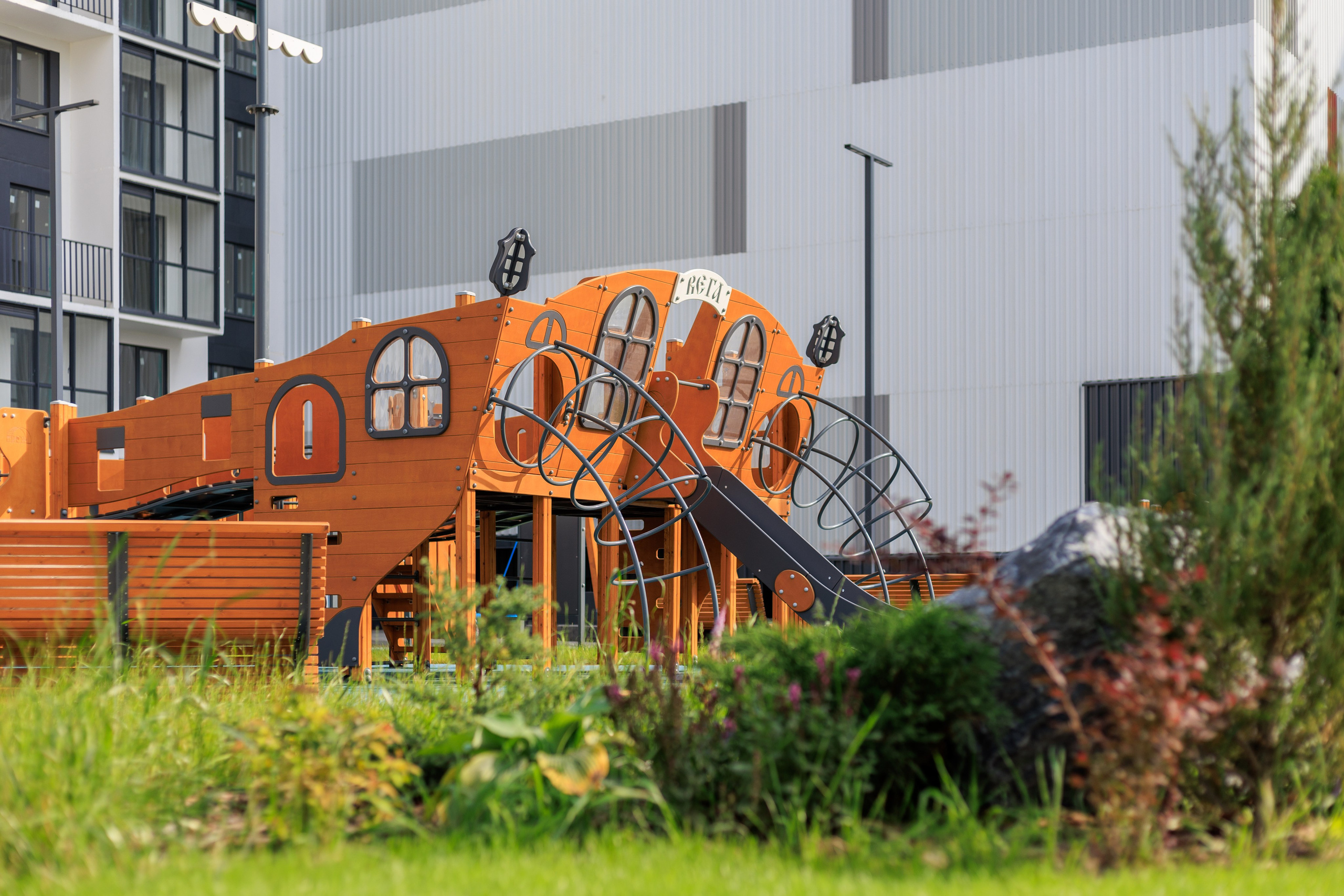 На изображении видна часть здания с ярко выраженной архитектурой, которая включает большие окна и необычные формы. Вокруг здания расположена зеленая зона с растениями и кустарниками.