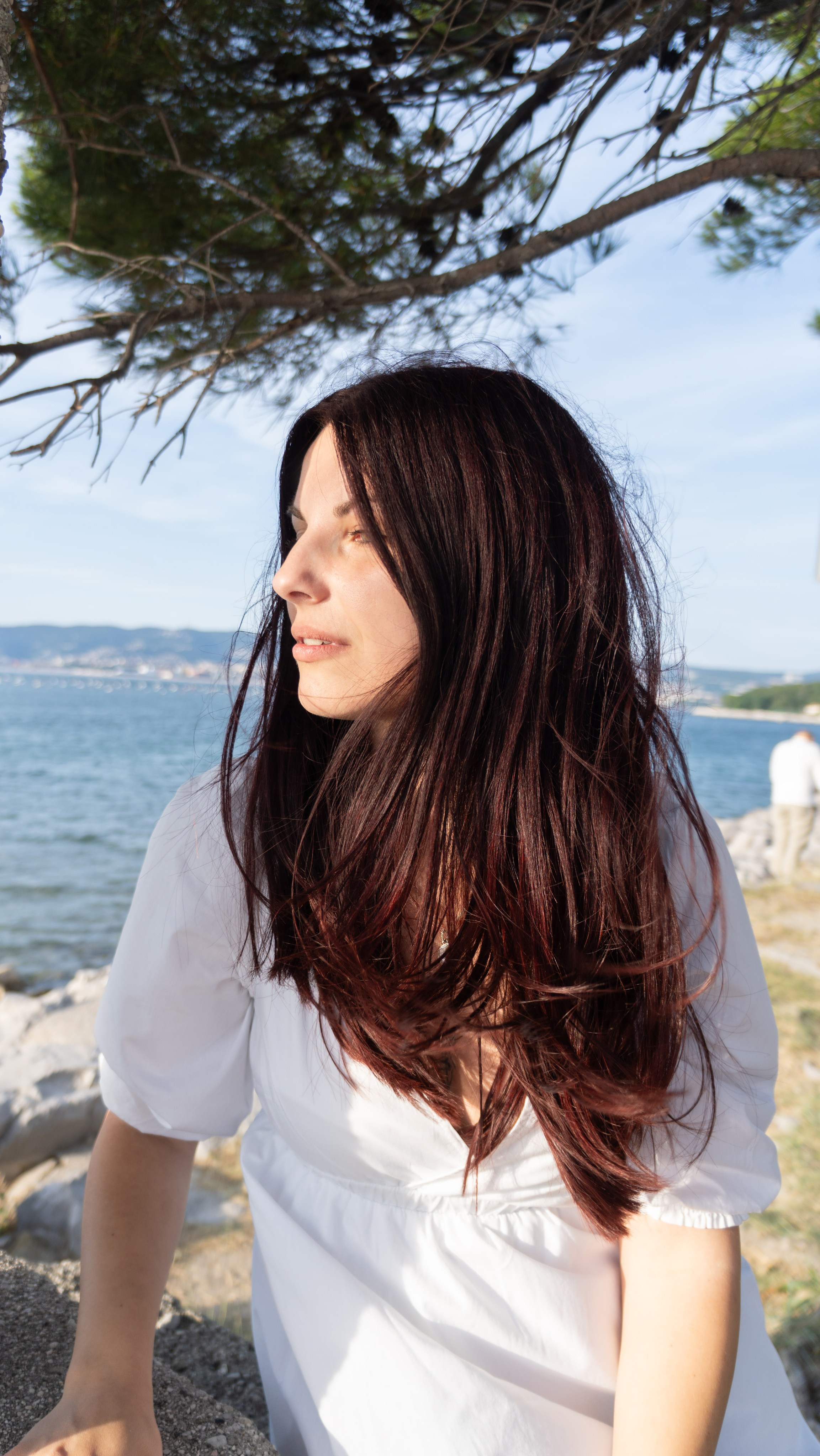 Ritratto di donna con lunghi capelli castani al vento, in riva al mare sotto la luce naturale.