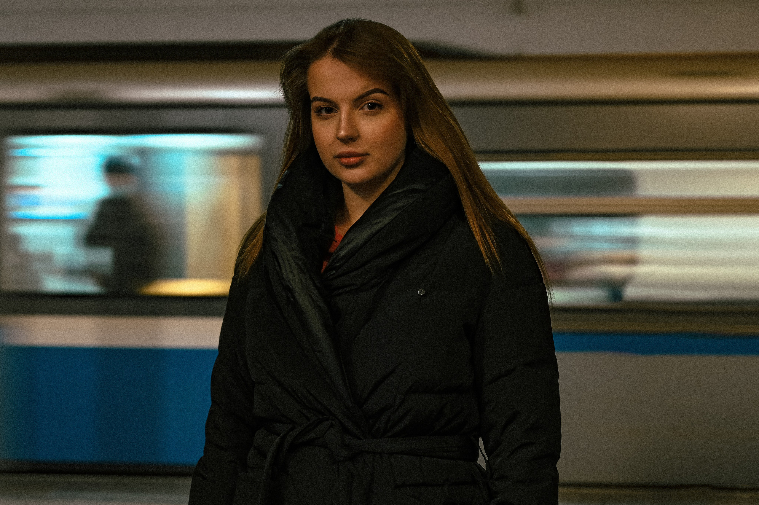 Молодая женщина стоит на платформе станции метро. Она одета в темный пуховик и имеет длинные волосы, которые свободно спадают по плечам. В фоне виден движущийся поезд, который создает эффект размытия, подчеркивая динамику городской среды. Освещение станции яркое, что делает образ модели четким и выразительным. Женщина смотрит прямо в камеру, демонстрируя уверенность и сосредоточенность. Фотография передает атмосферу ожидания и спокойствия в условиях активного движения вокруг нее.