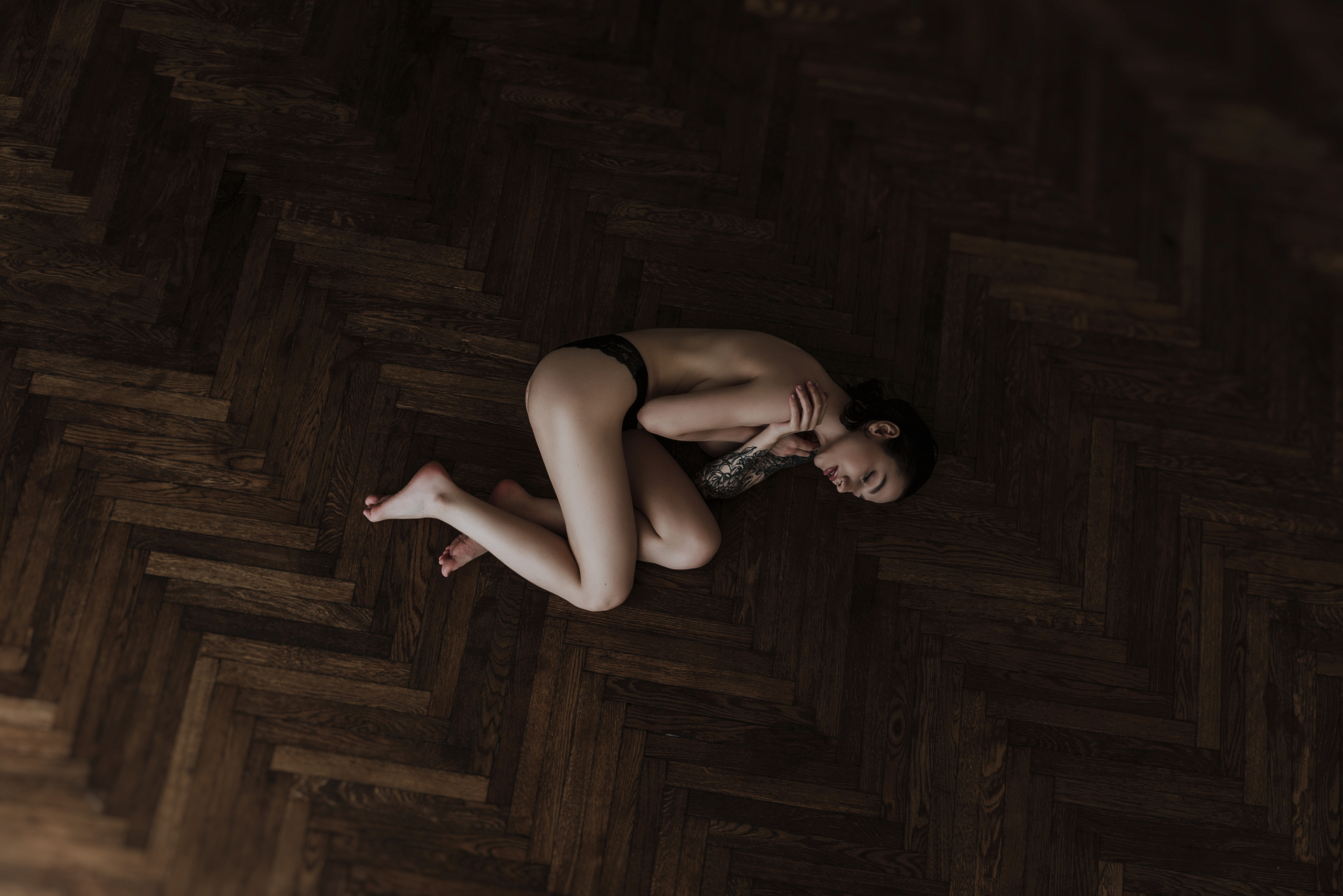 Девушка лежит в позе эмбриона на коричневом паркете.