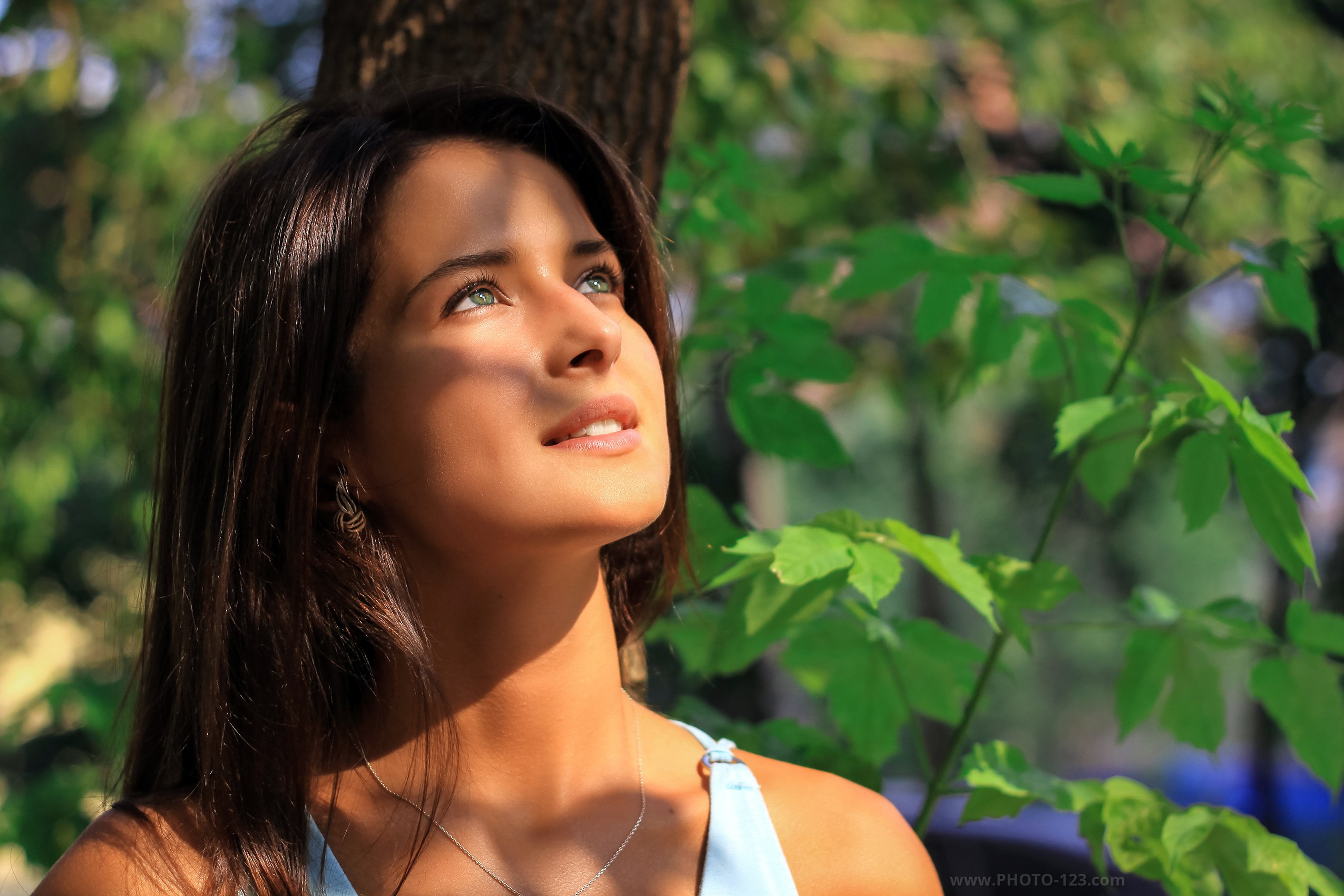 Young woman standing in a forest, looking upward toward sunlight filtering through green leaves, soft natural light casting gentle shadows on her face, calm and reflective expression, outdoor portrait photography, natural beauty, serene woodland atmosphere