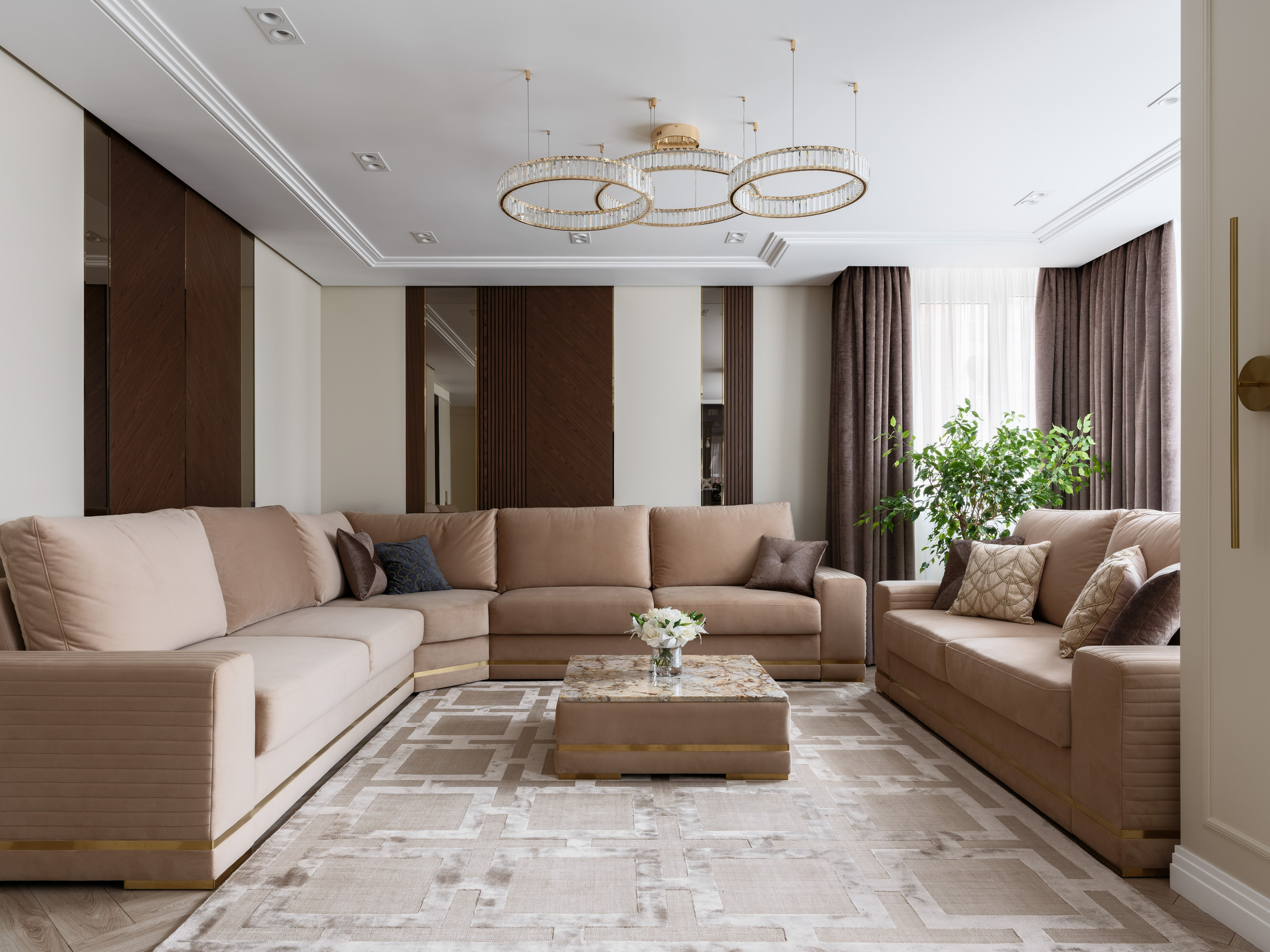 Современная гостиная в стиле неоклассика с тремя мягкими диванами в бежевой обивке, журнальным столиком с каменной столешницей и ковром светлого оттенка с геометрическим узором, дополненная шторами и зелёным растением.