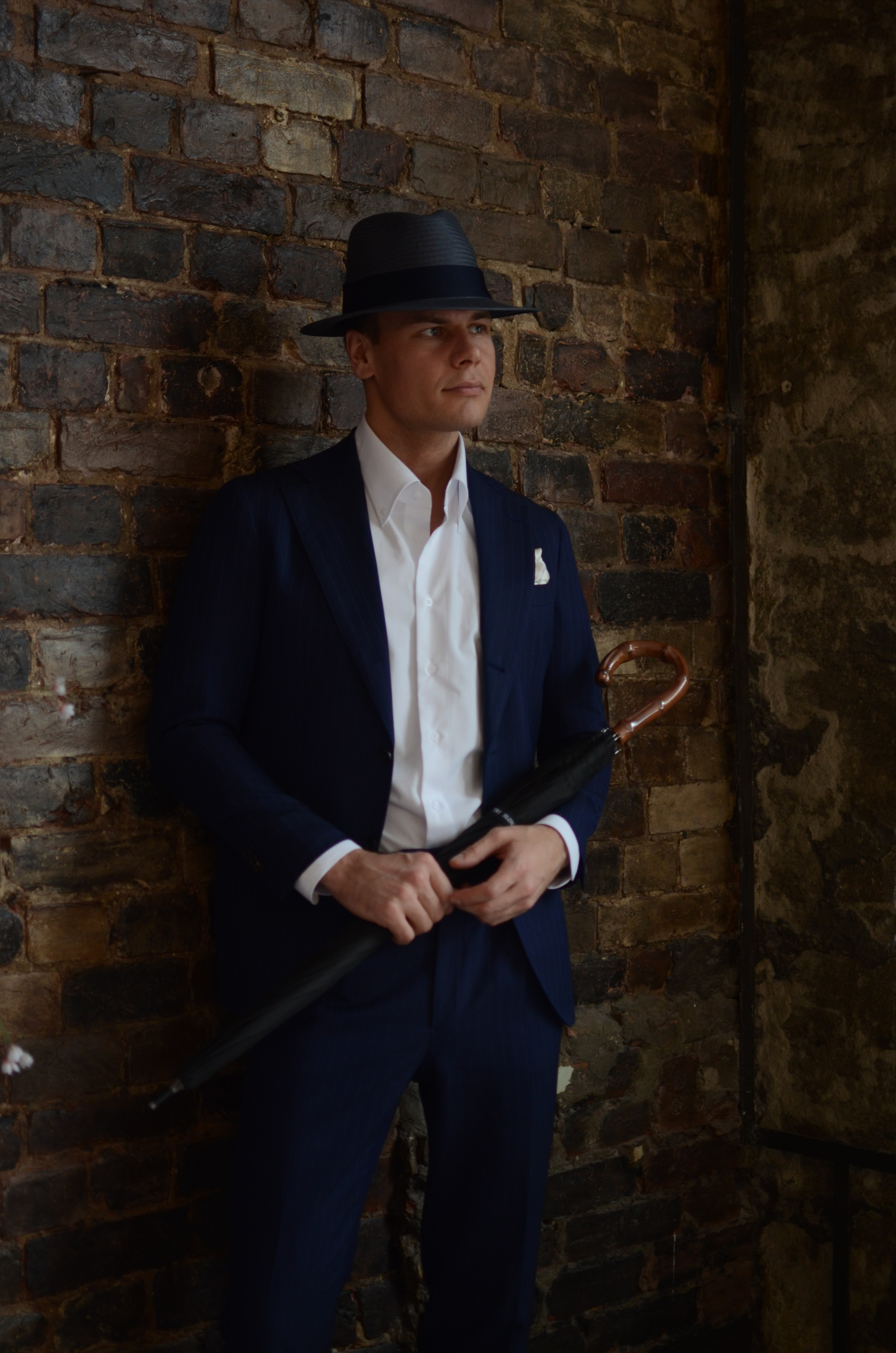 Мужчина в деловом костюме в шляпе и зонтов руках стоит у кирпичной стены