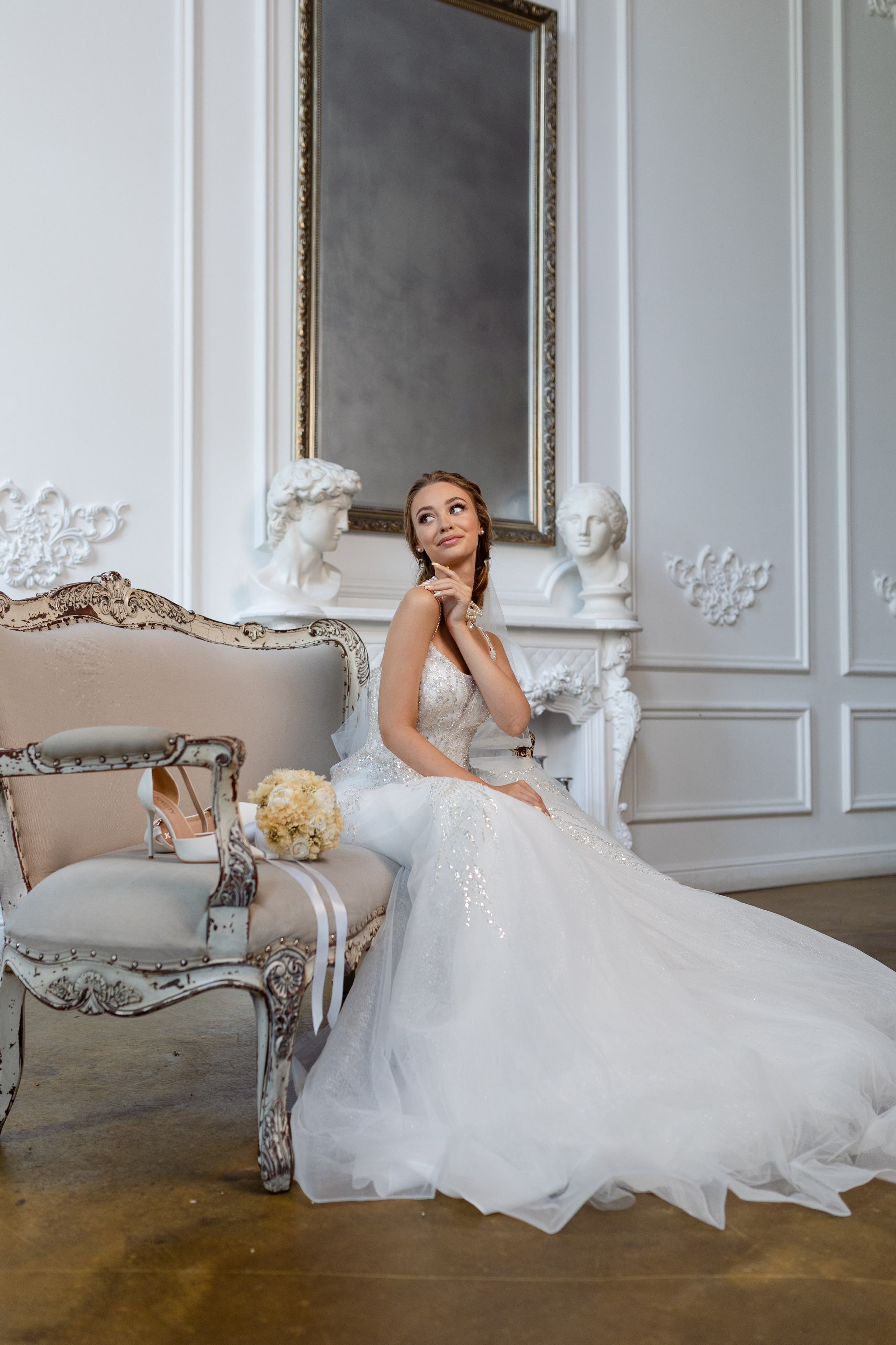 Невеста сидит на диване, на котором лежат туфли невесты и ее букет.