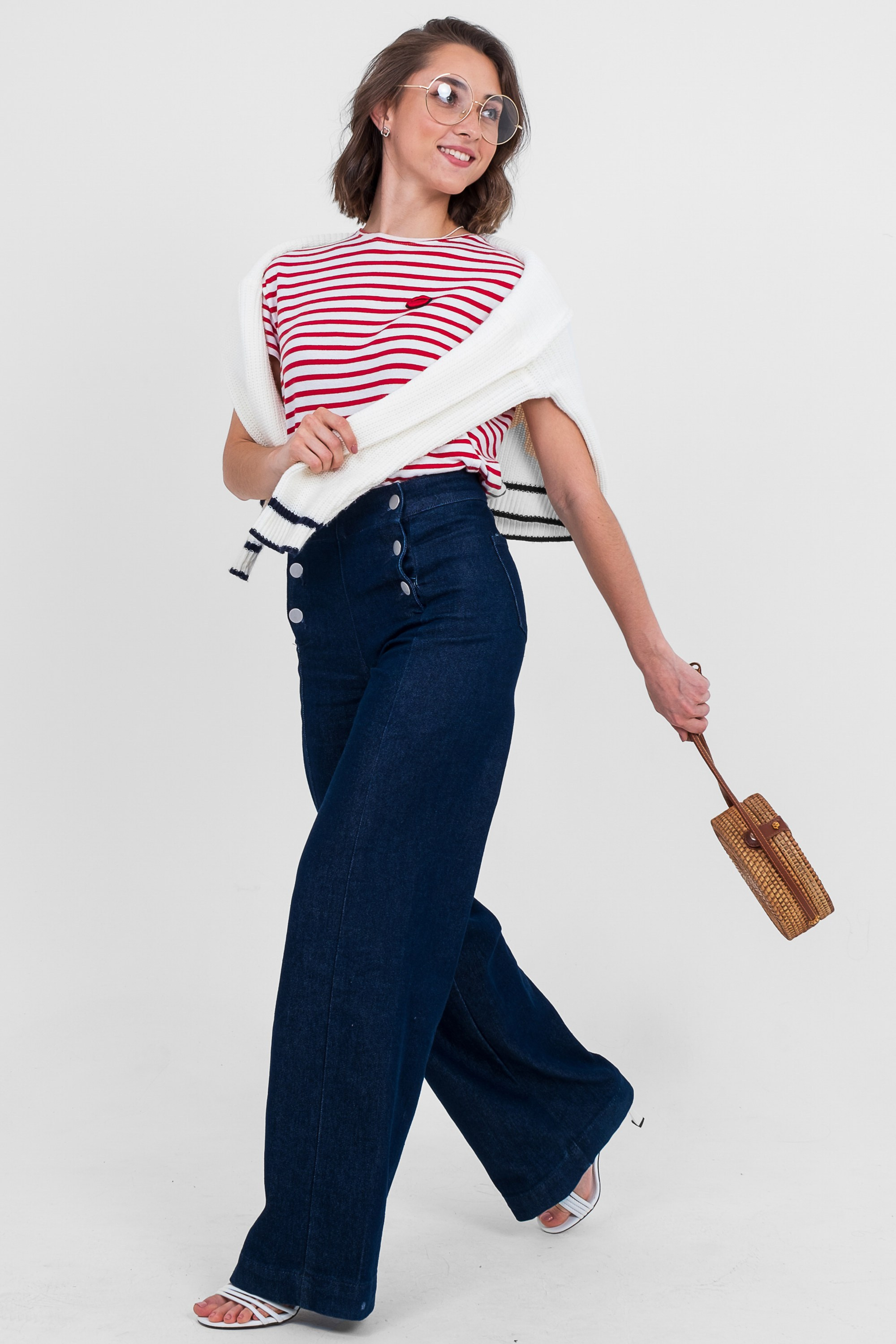 На изображении девушка в очках в полосатой футболке с белой кофтой и широких джинсах с пуговицами и с сумкой.  Фото для маркетплейсов