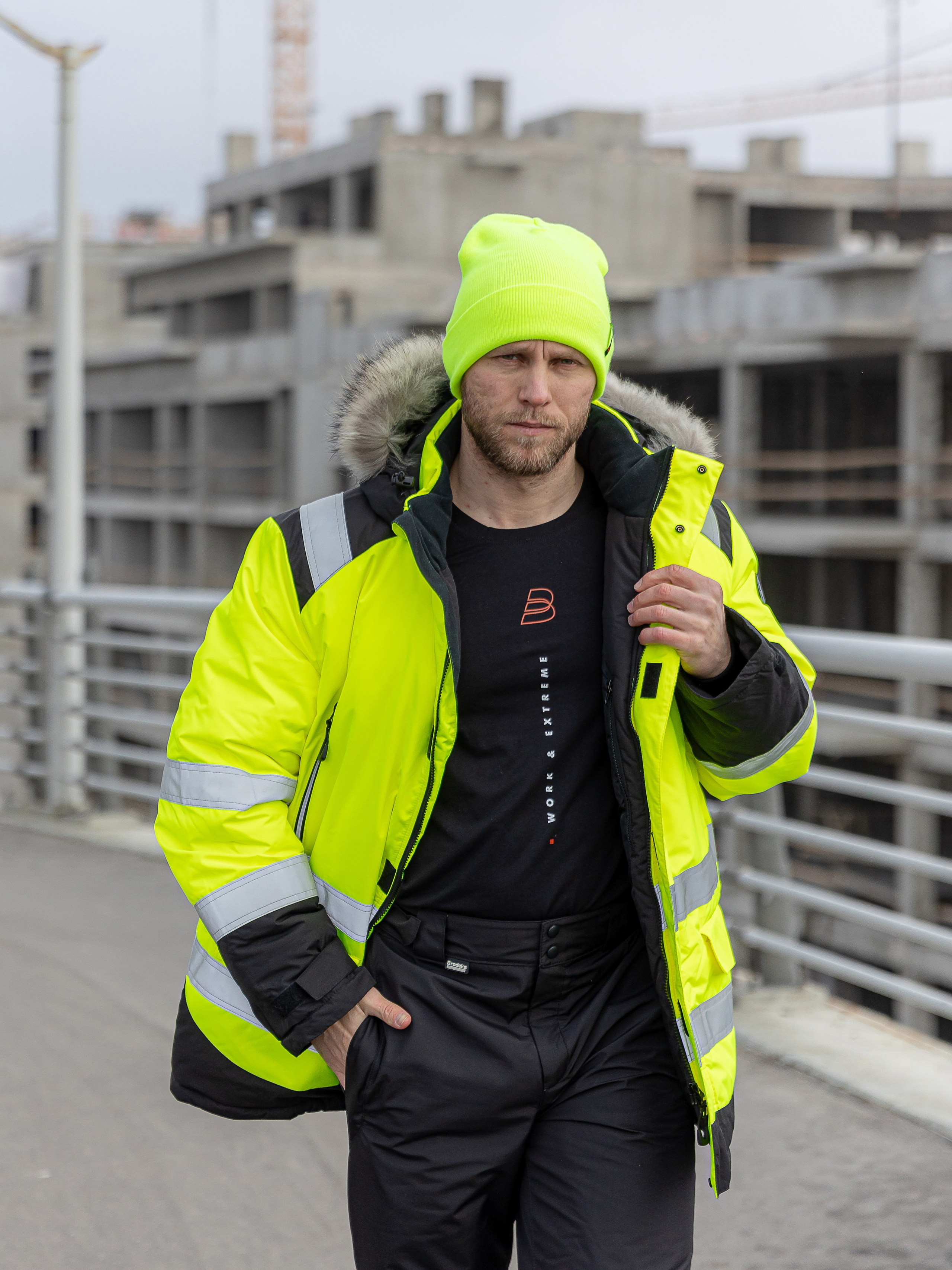 Брутальный мужчина в сигнальной куртке стоит на фоне стройки