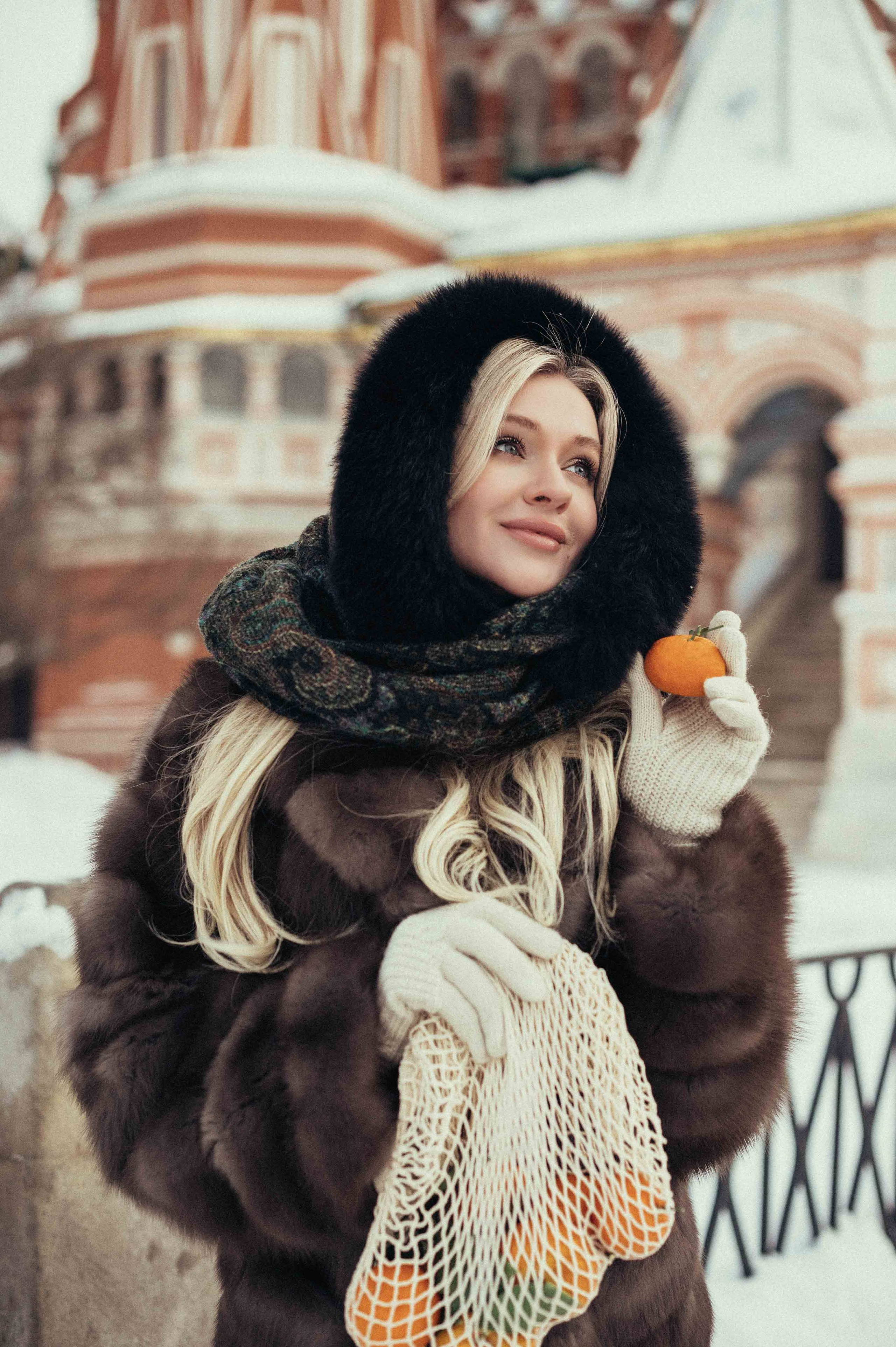 Девушка в стиле Russian girl  крупным планом на фоне храма Василия Блаженного на Красной площади в Москве, в правой руке которой авоська с мандаринами, а в левой один мандарин