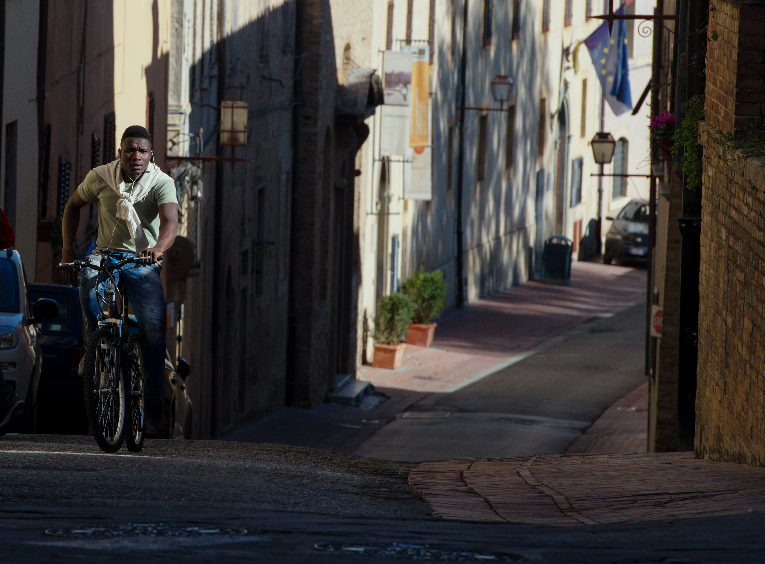 Стрит-фотография мужчины на велосипеде, поднимающегося по узкой, залитой солнцем улице старого европейского города.