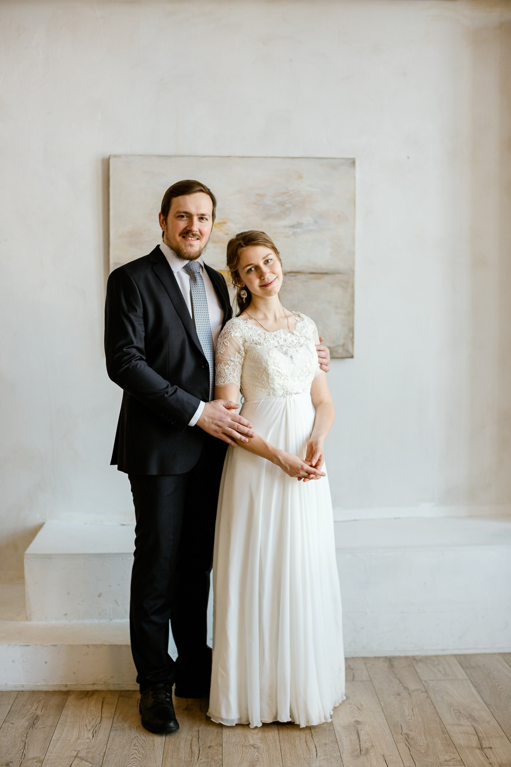 Жених и невеста в праздничной фотостудии. Жених в костюме, невеста в красивом белом платье. На фоне картина.