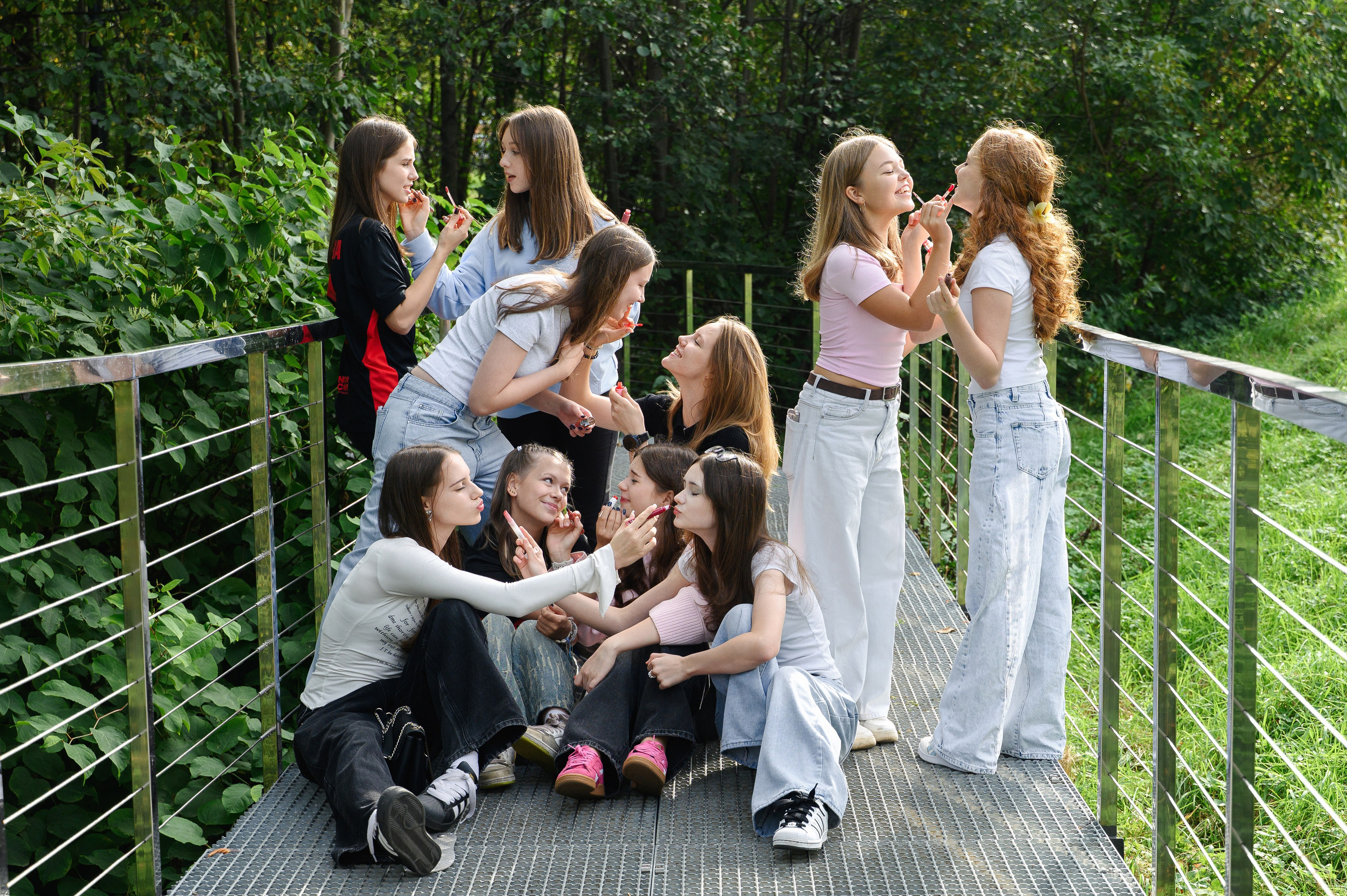 Группа выпускниц красит друг другу губы на подвесном мосту на фоне густой летней зелени.