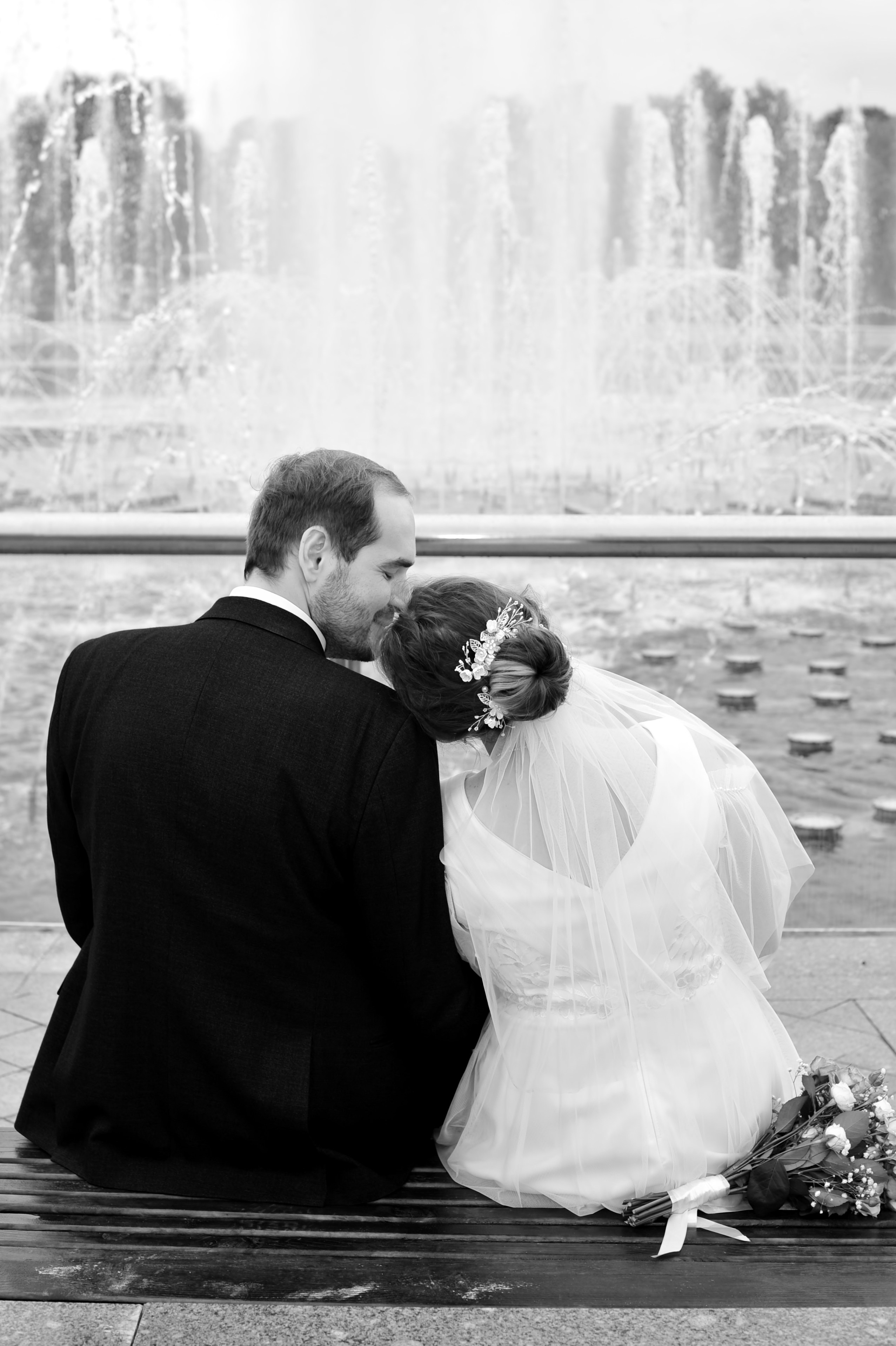 Свадьба в Царицыно пасмурным летним днем, новобрачные сидят на лавочке спиной к зрителю и лицом к фонтану, невеста положила голову на плече жениху,  черно-белая фотография