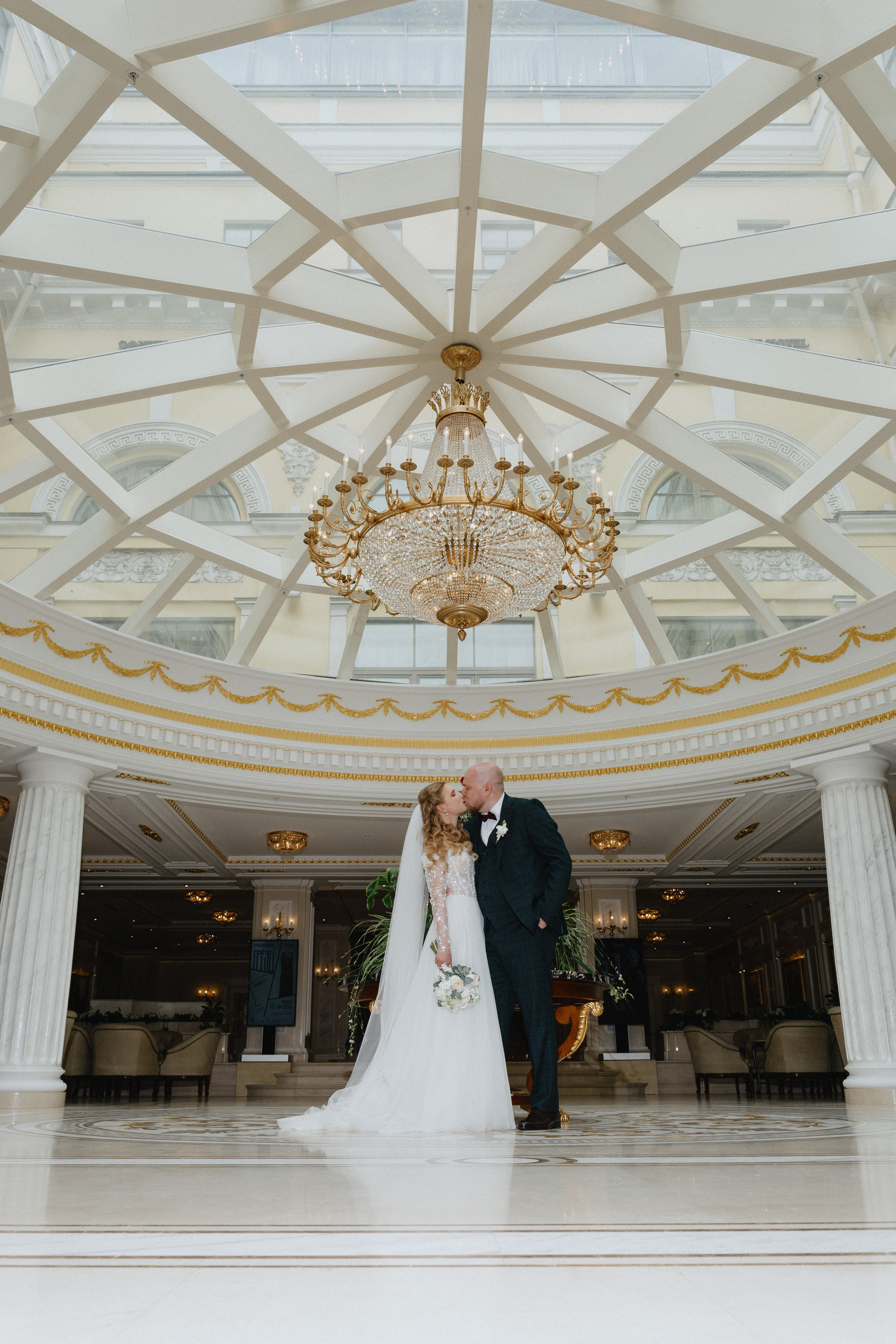 Свадебная фотосессия в роскошном интерьере отеля Эрмитаж: жених и невеста стоят под большим стеклянным куполом с люстрой.