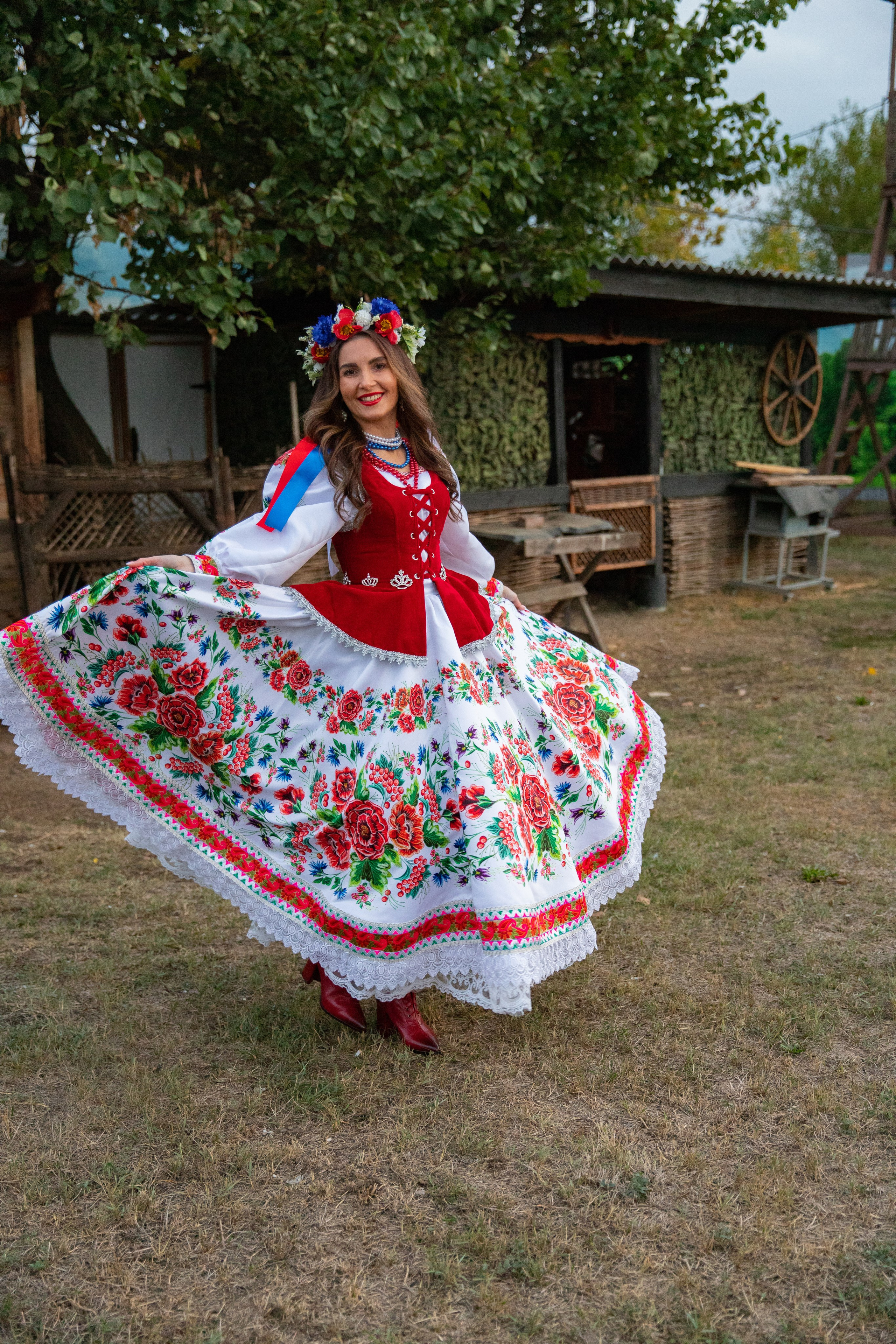 Изящное платье казачки эффектно развевается на ветру, девушка показывает его детали на фоне зелёных полей.