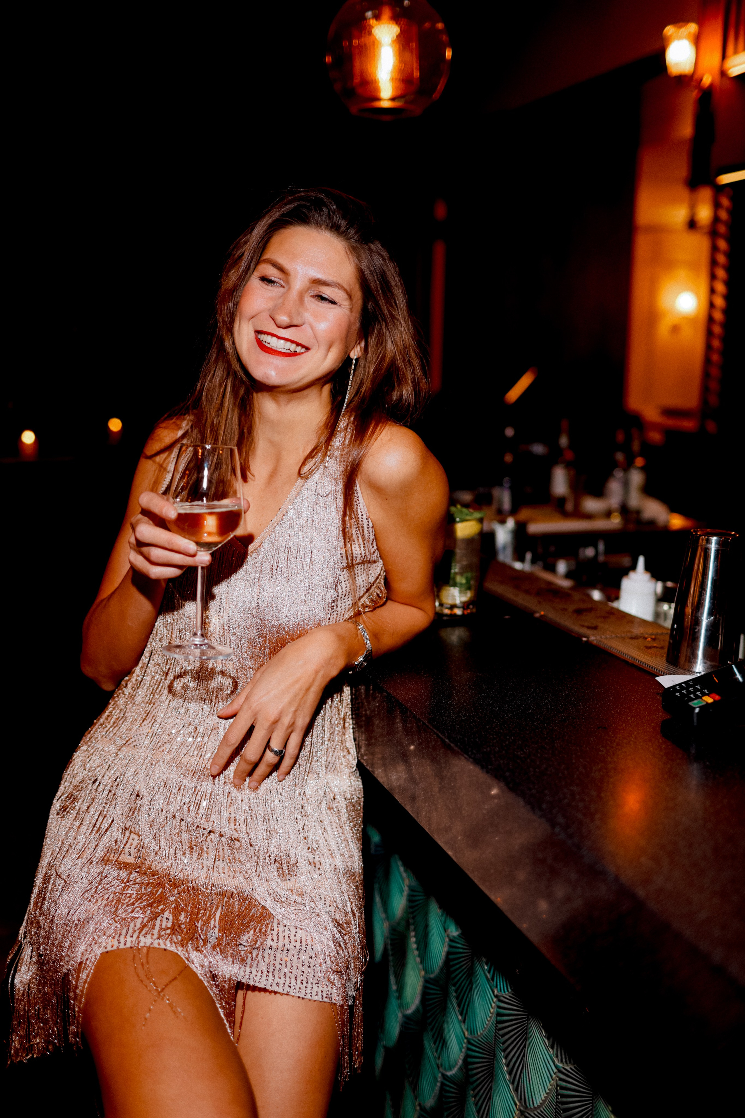 <img src="IZ0A0352.jpg" alt="Красивая девушка в блестящем платье с бахромой сидит в баре ночью  с бокалом в руке  широко улыбается ">