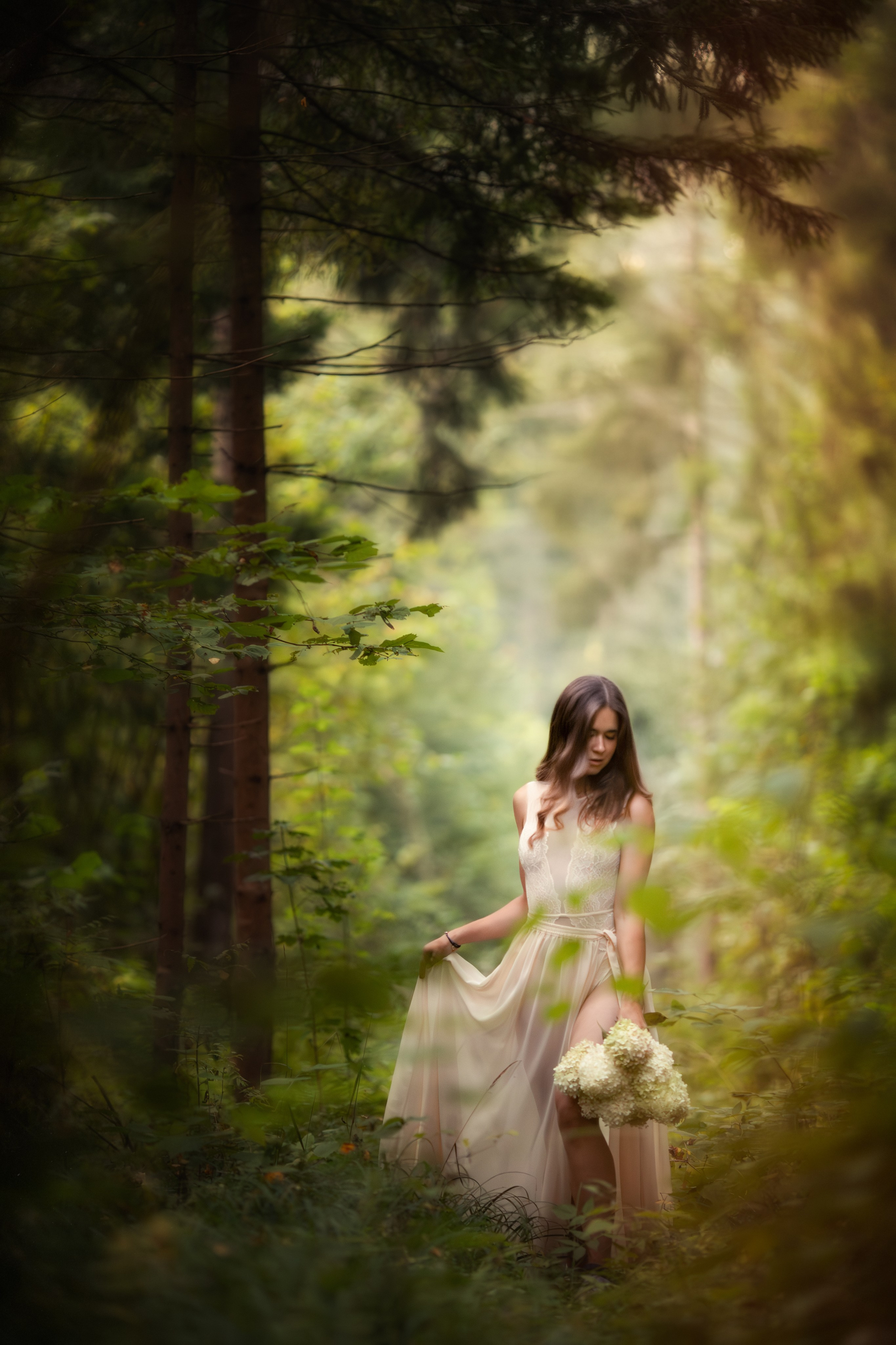 Девушка в длинном светлом платье стоит в лесу с букетом белых цветов, вокруг густая зелень и мягкий свет, создающий атмосферу тишины и уюта.