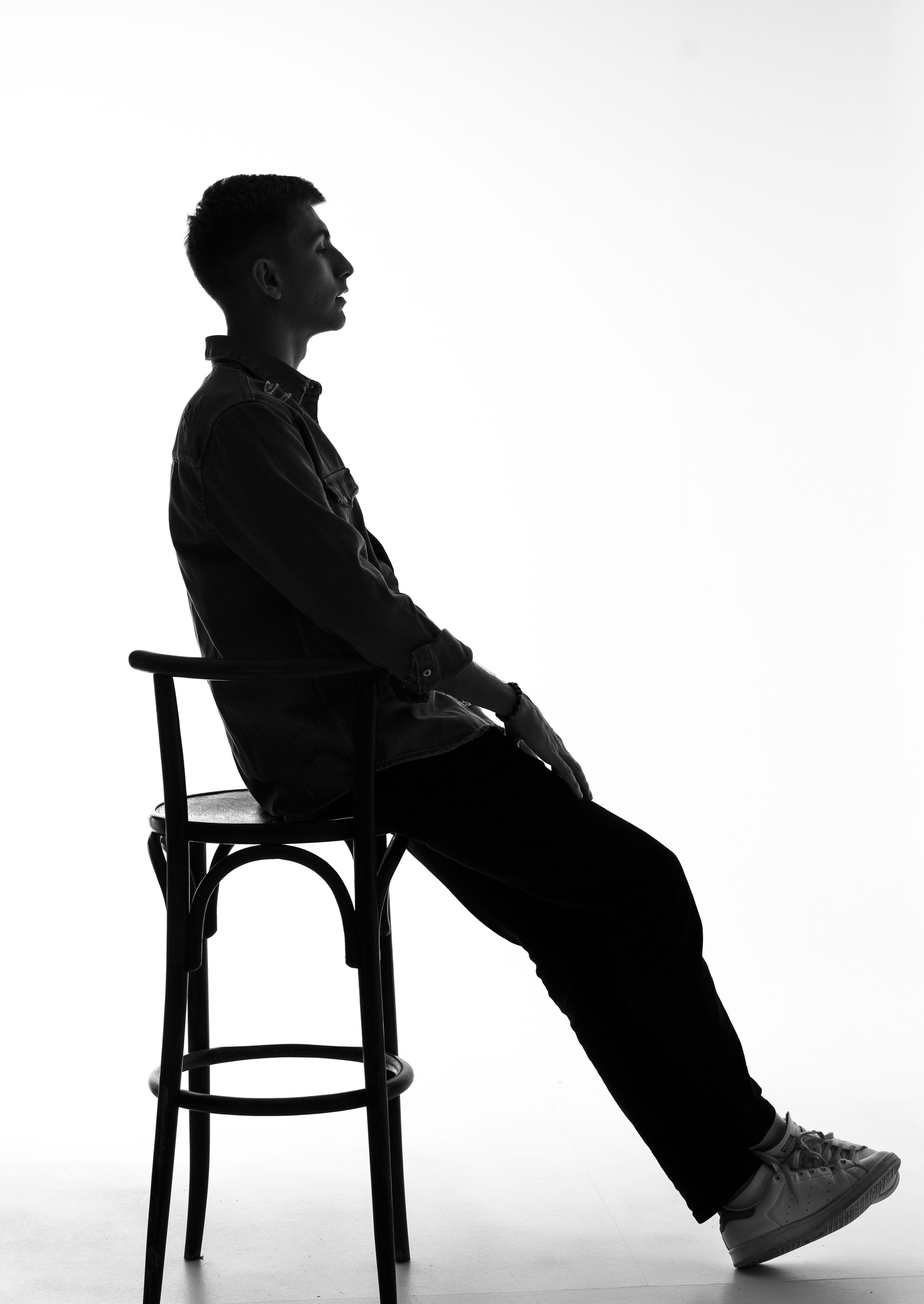 На фотографии изображен человек сидящий на стуле. Он, по-видимому, одет в рубашку с пуговицами и брюки, а также обут в туфли. Фотография сделана на белом фоне, и освещение создает силуэтный эффект на этом человеке.