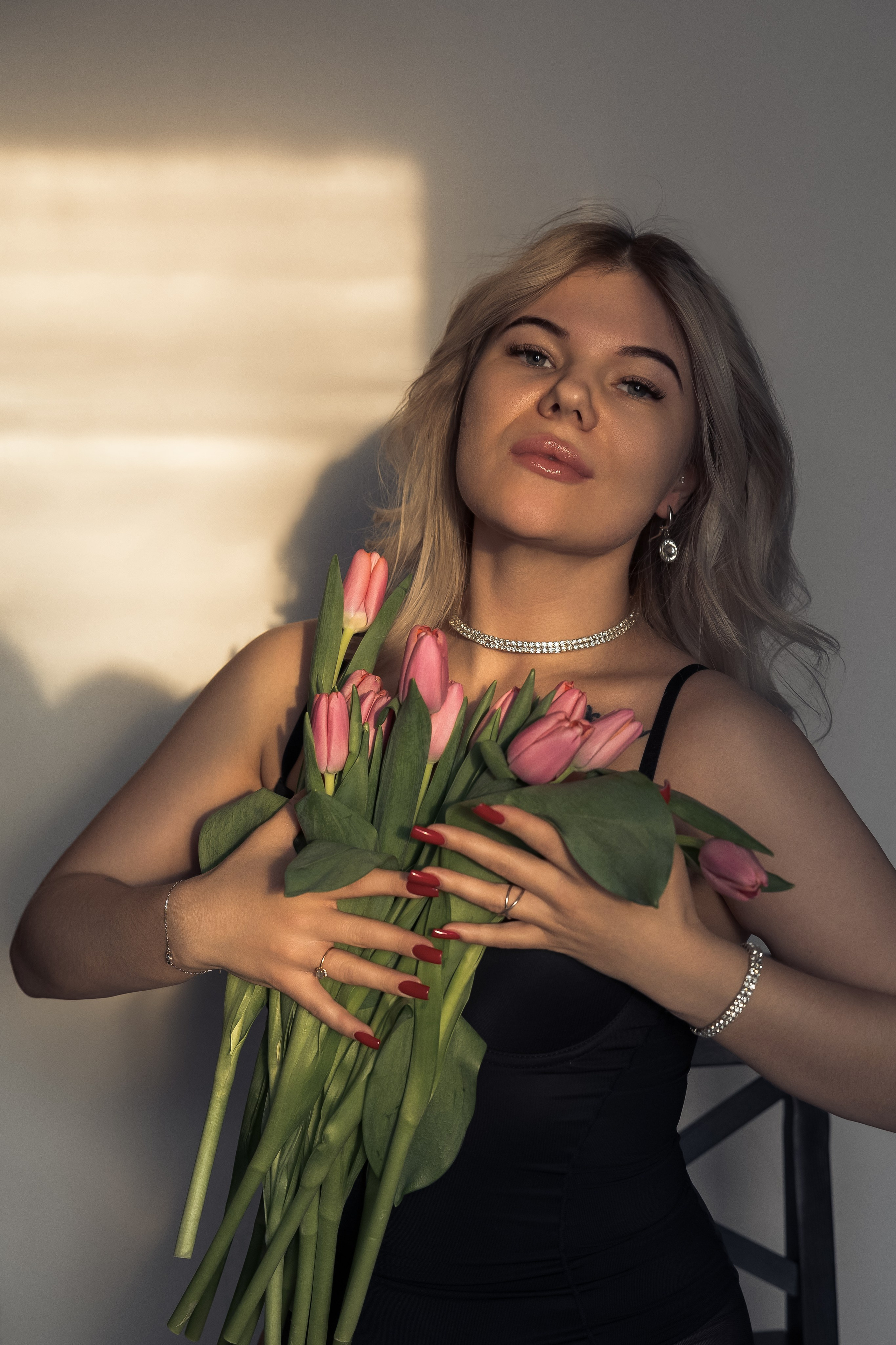 Девушка стоит у стены в закатном свете, держит букет тюльпанов и улыбается.