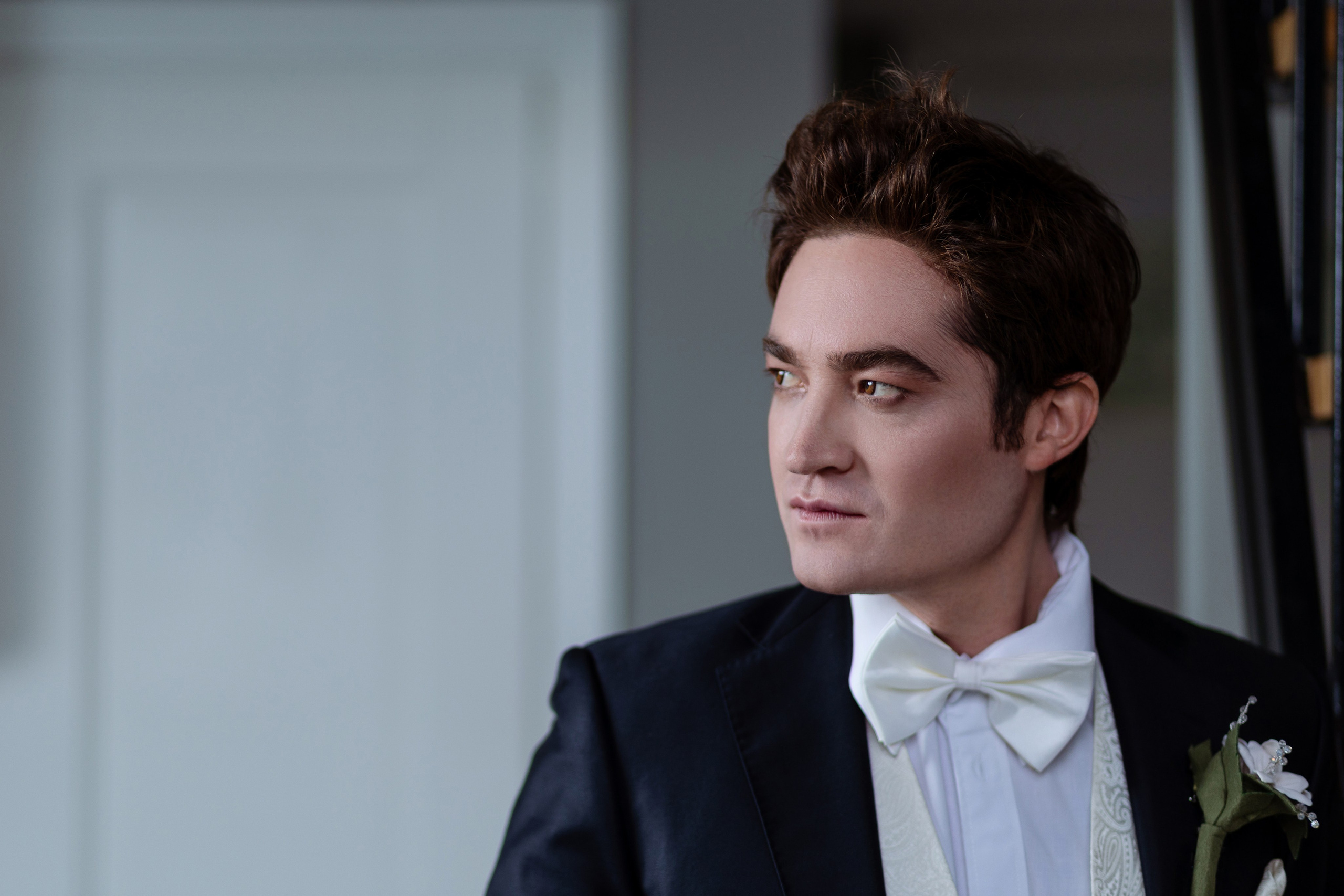 Жених стоит на фоне белой стены в полупрофиль.