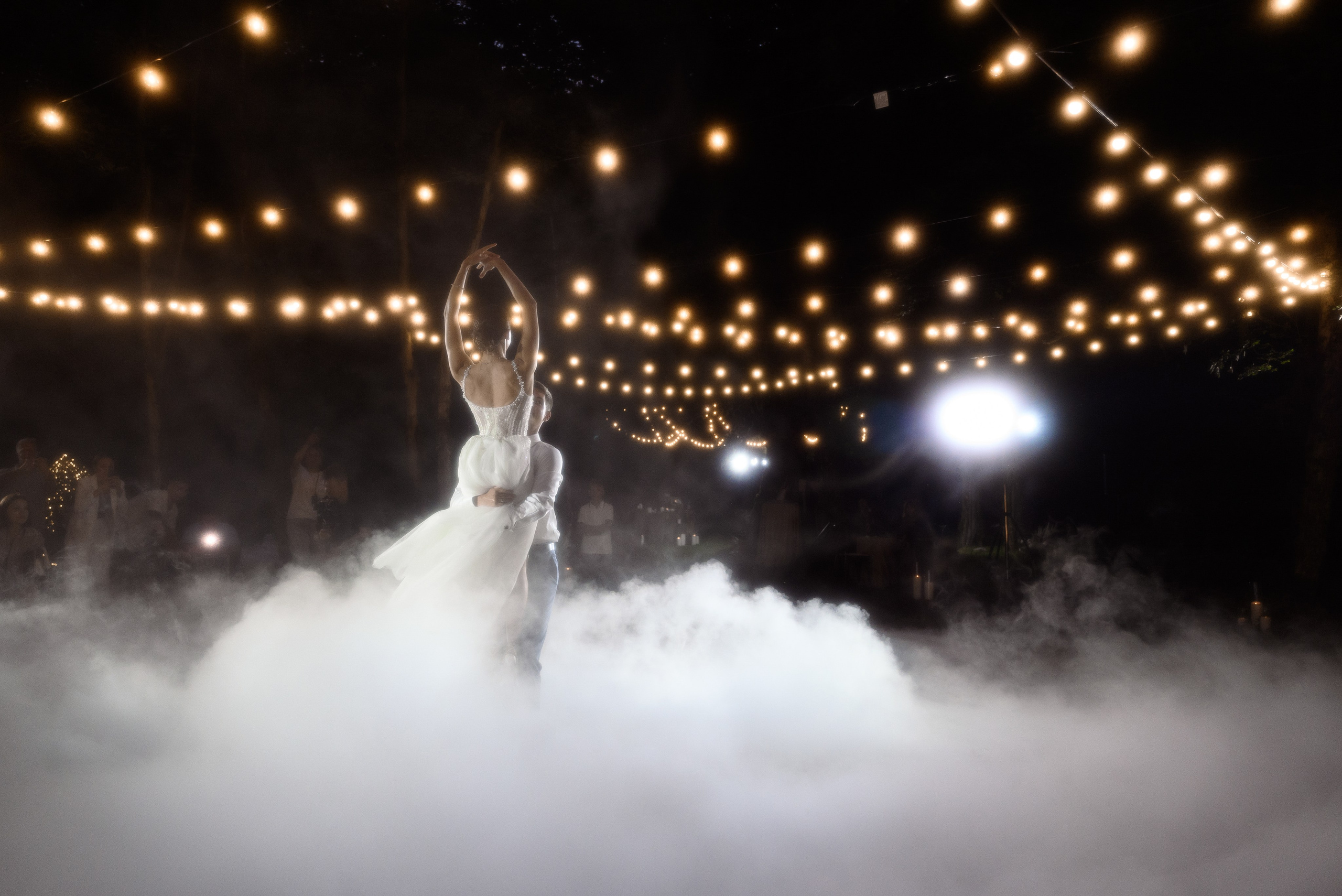 Свадебные фотографии в Сочи, в глэмпинге Енот среди природы, где любая пара может найти гармонию и романтику на свадьбе они танцуют свой первый танец вокруг темно гирлянда сверху и всё заполнено снизу кадра густым густым дымом 