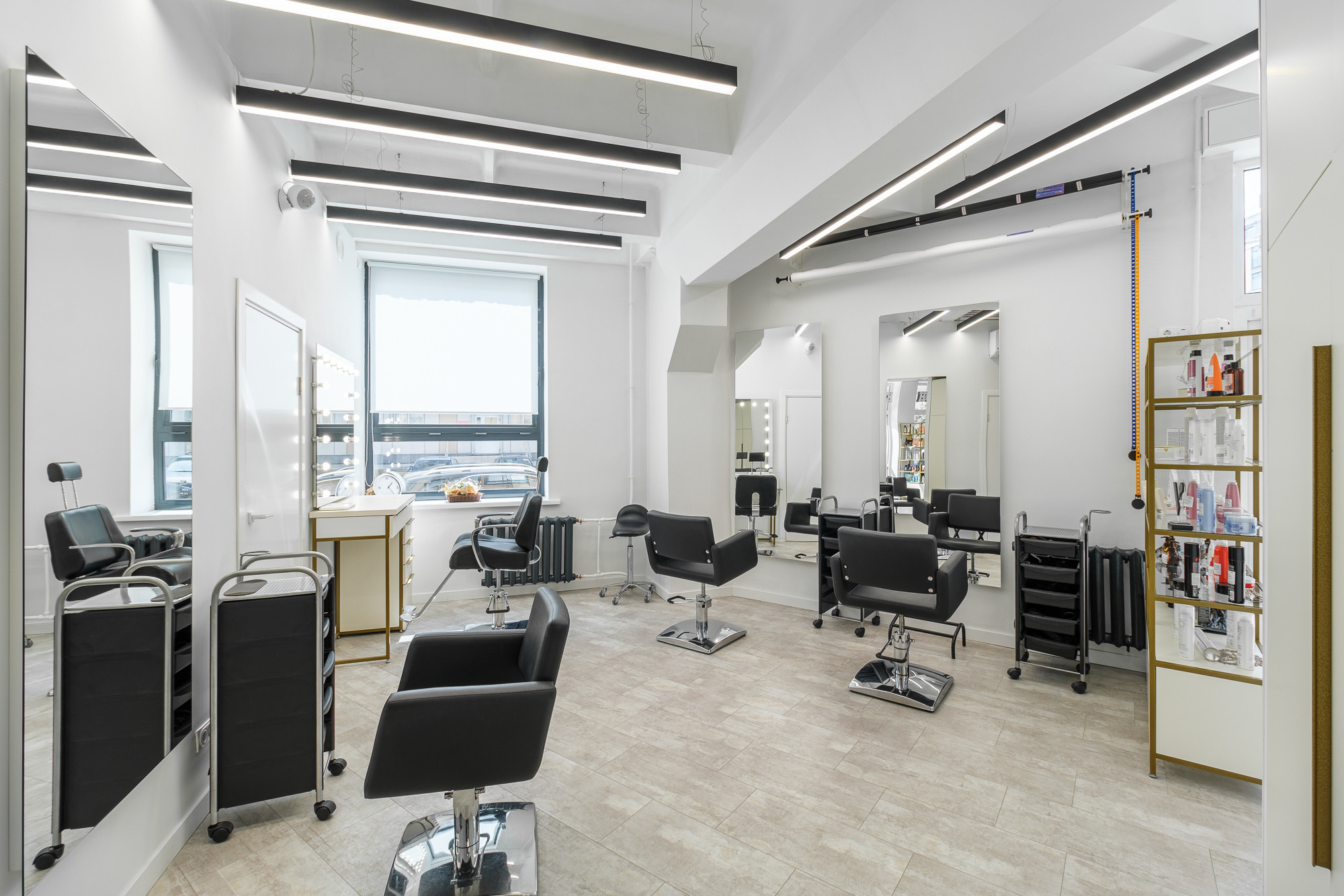 На изображении представлен салон красоты с современным интерьером. Видно несколько рабочих мест для парикмахеров, оборудованных креслами и зеркалами. Салон имеет хорошее освещение и просторное помещение.
