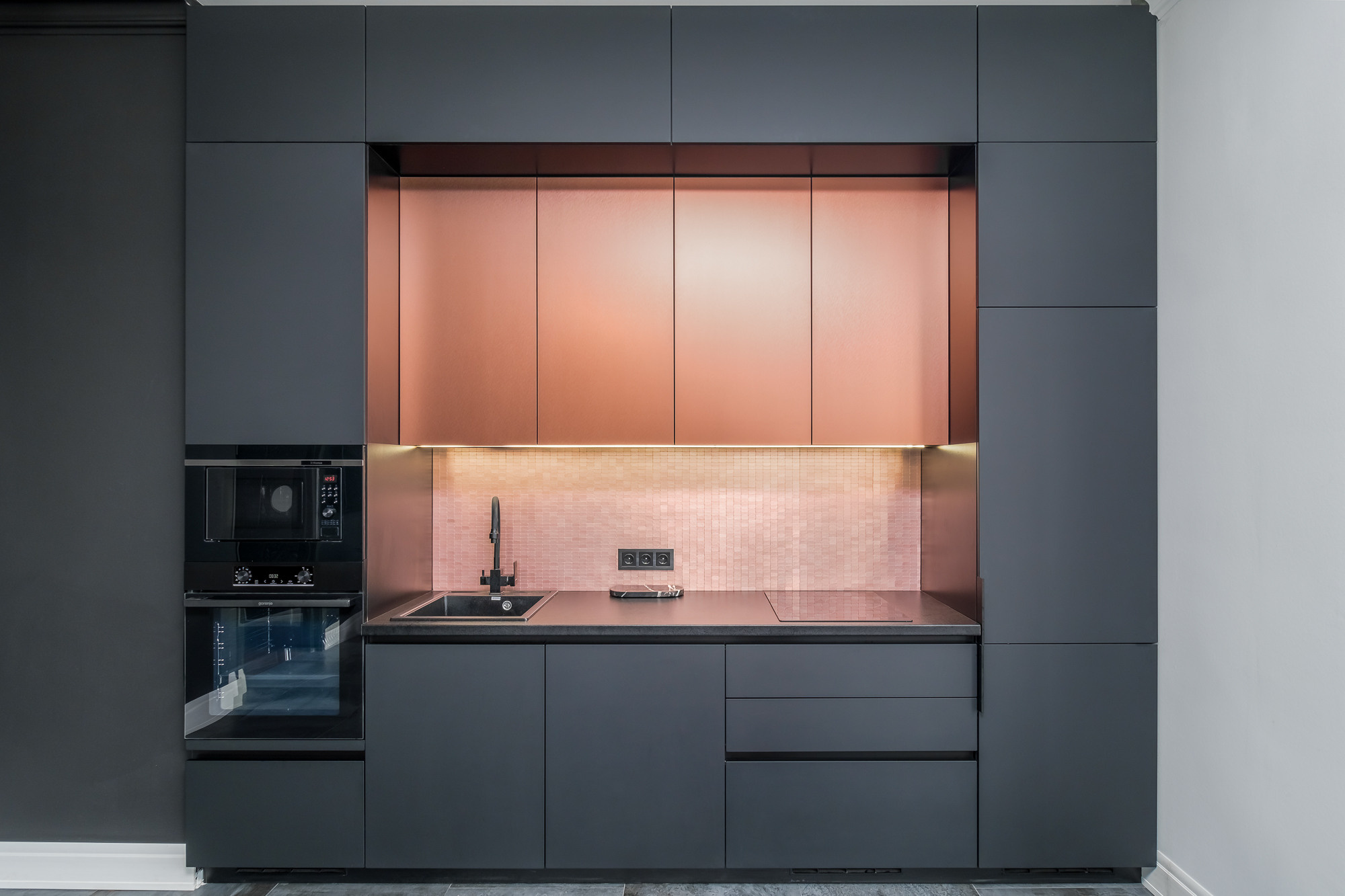 На изображении представлена современная кухня с минималистичным дизайном. Видна столешница темного цвета, кухонные шкафы и бытовая техника. Акцентом кухни является фартук медного цвета.