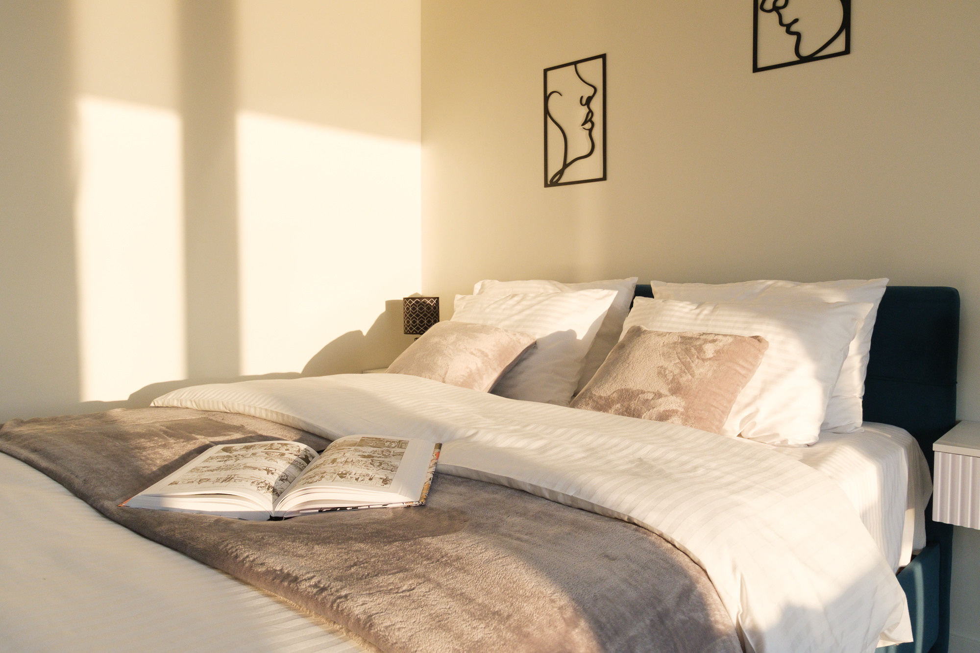 На изображении спальня c аккуратно застеленными постельным бельем. На кровати лежит книга. В комнате много закатного света, поступающего через большое окно, и на стенах висят картины.