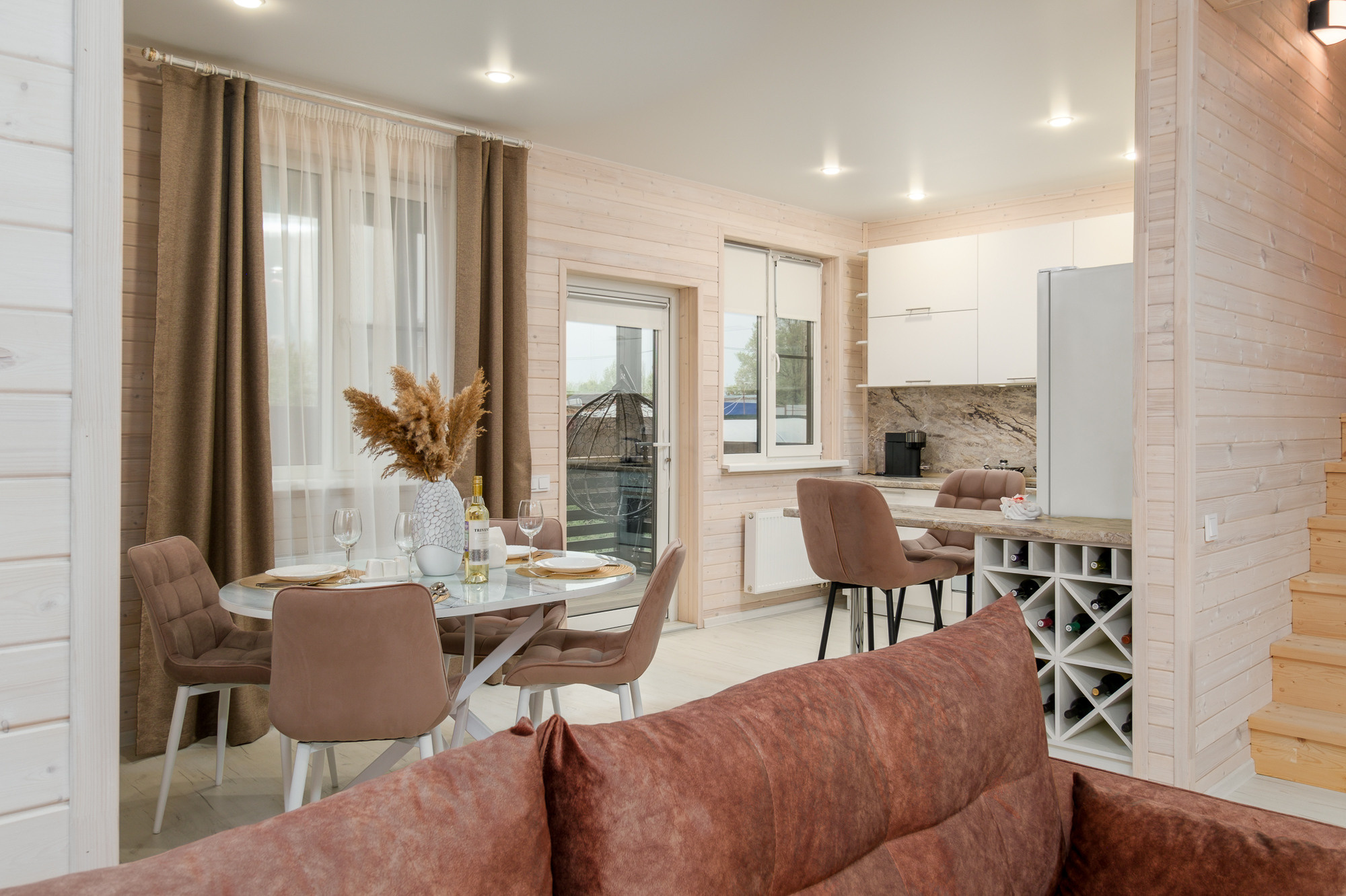 Гостиная с диваном коричневого цвета, столом и стульями для столовой зоны. В комнате также есть большие окна с шторами и выход на террасу.