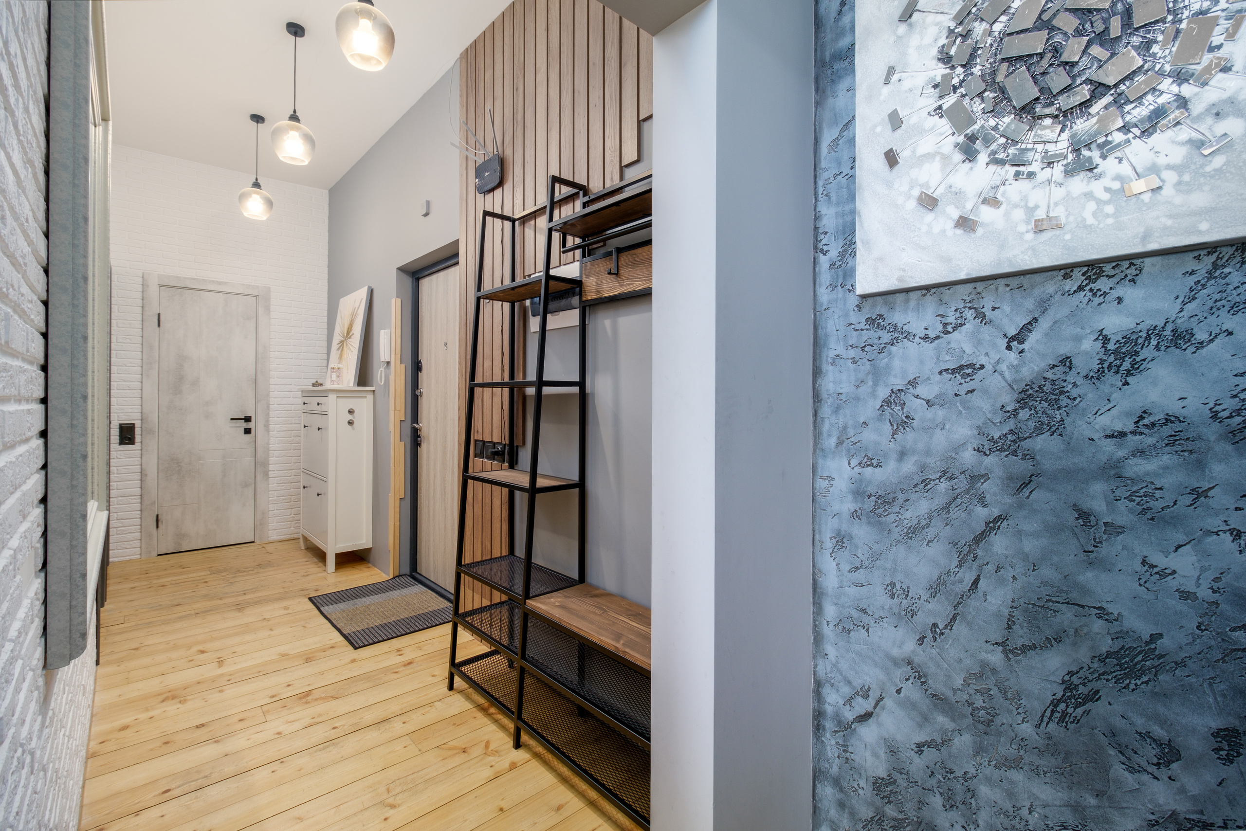 На изображении показан коридор квартиры с современным ремонтом. На переднем плане видна картина, на среднем плане находится стеллаж в лофт-ст иле, за ним белый комод. Стены вокруг окрашены в серый цвет с различными оттенками и текстурами.