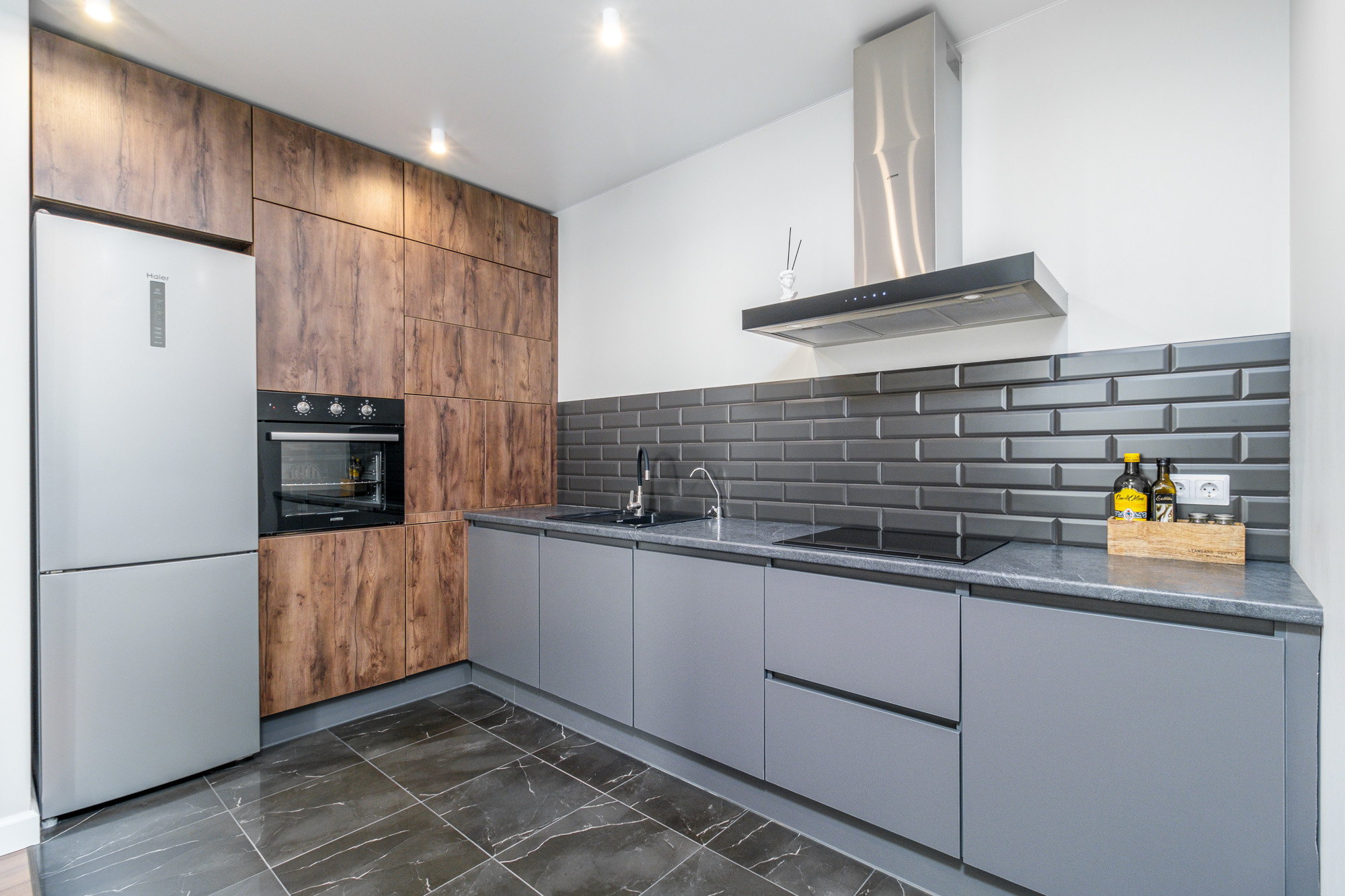 Современная кухня с серой мебелью и деревянными элементами. В центре расположена плита с черной варочной поверхностью, а над ней висит вытяжка. Слева находится встроенный холодильник серебристого цвета. Пол выполнен из тёмной плитки с узором "под мрамор".