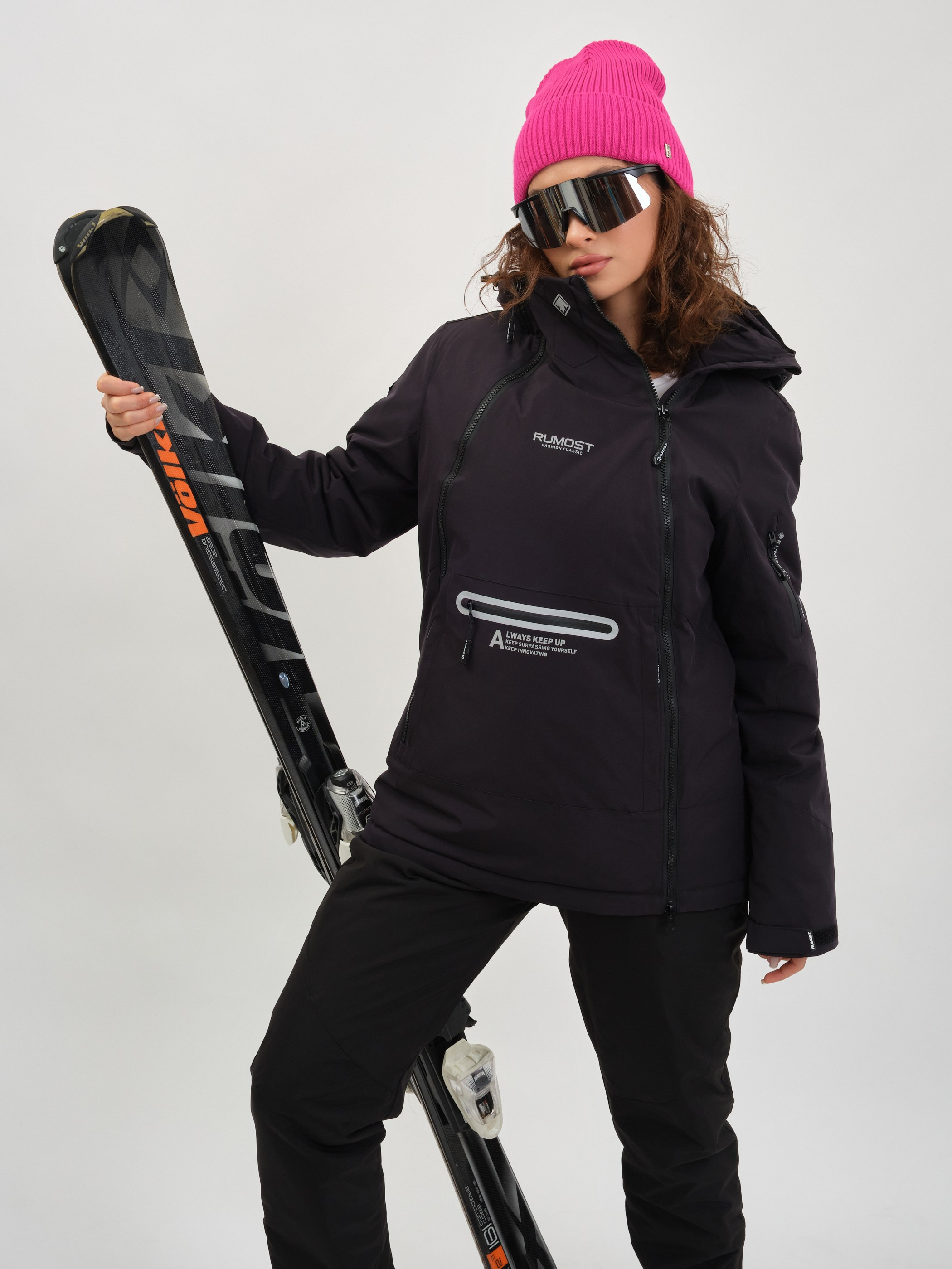 Девушка в зимней лыжной одежде в спортивных очках и розовой шапке стоит с лыжами