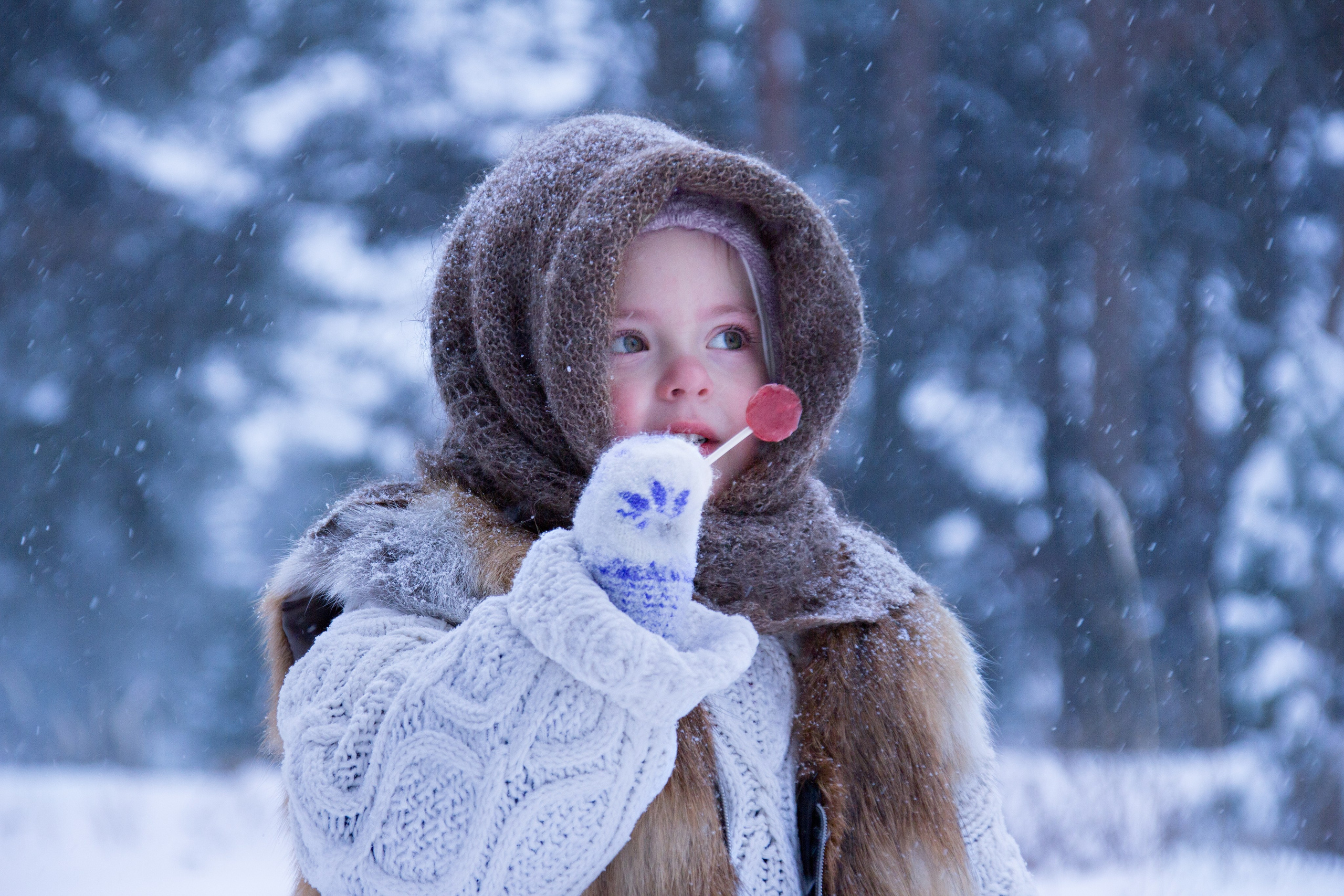 Девочка в пуховом коричневом платке и меховой жилетке ест леденец в снежном лесу