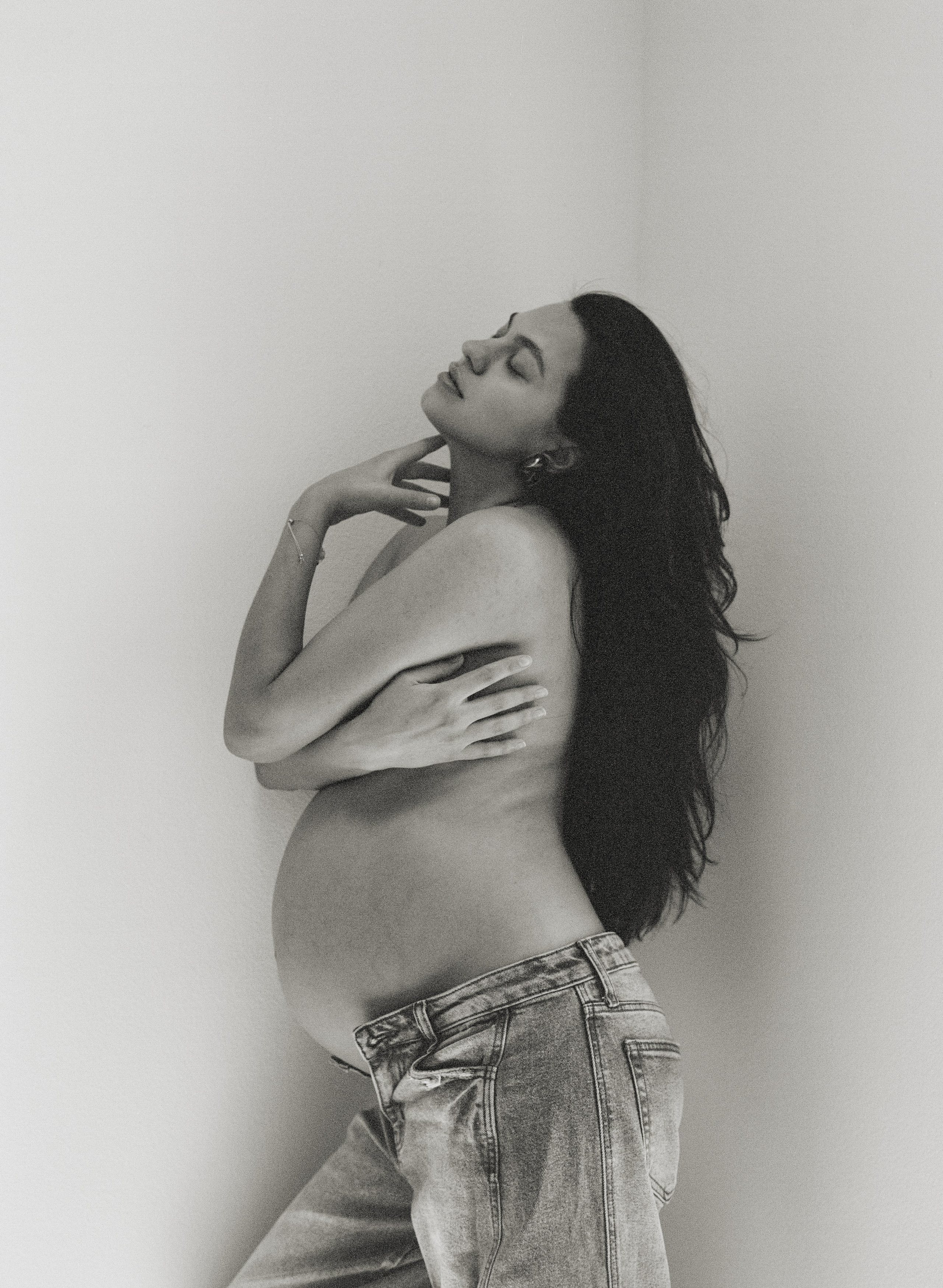 a black and white film photo of a pregnant woman in jeans covering top part of her body with hands and long dark hair