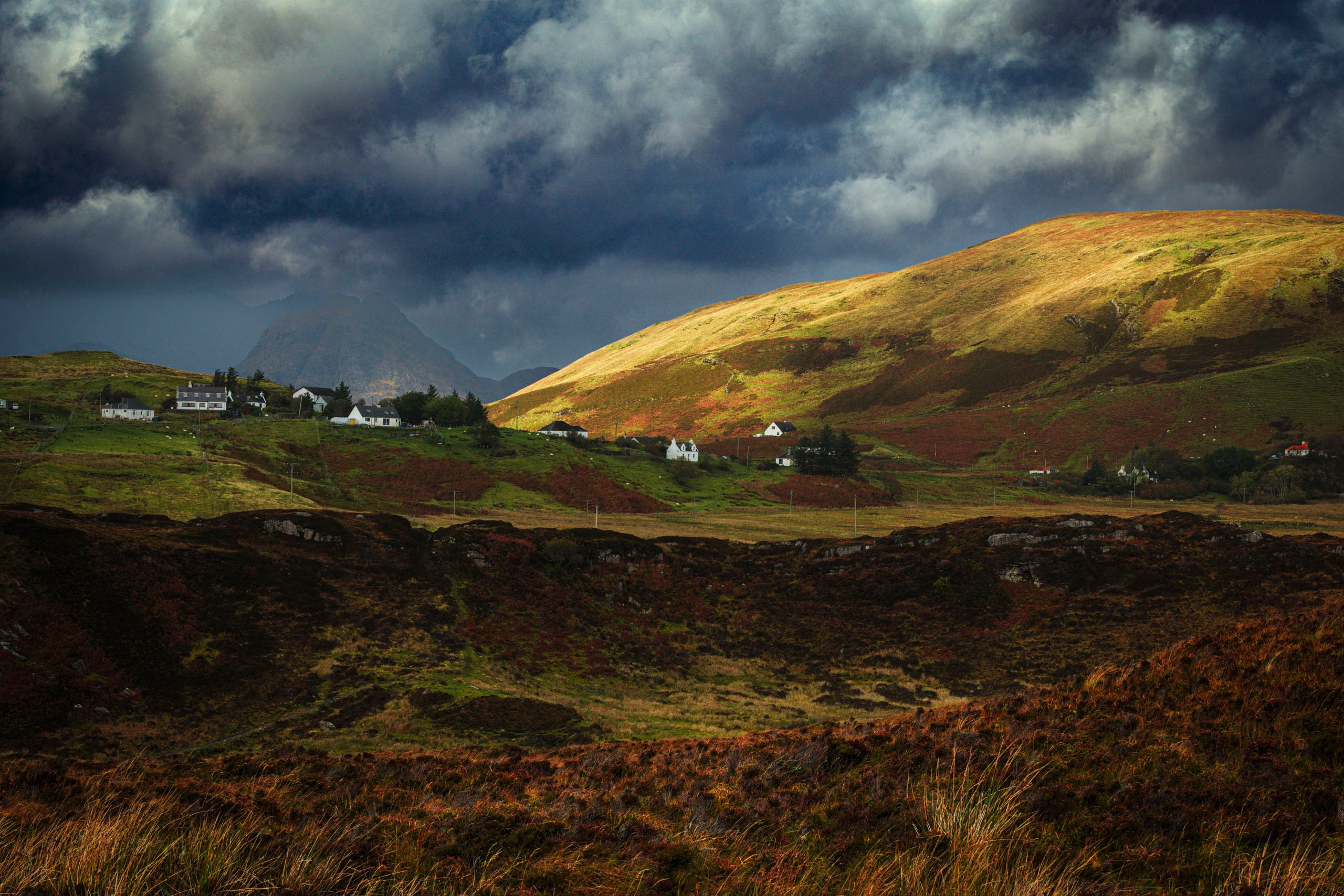 Атмосферный пейзаж в Шотландии: группа коттеджей и домов на зеленом склоне холма, освещенных солнечным светом сквозь драматичные темные облака, на фоне горного массива и вересковых пустошей.