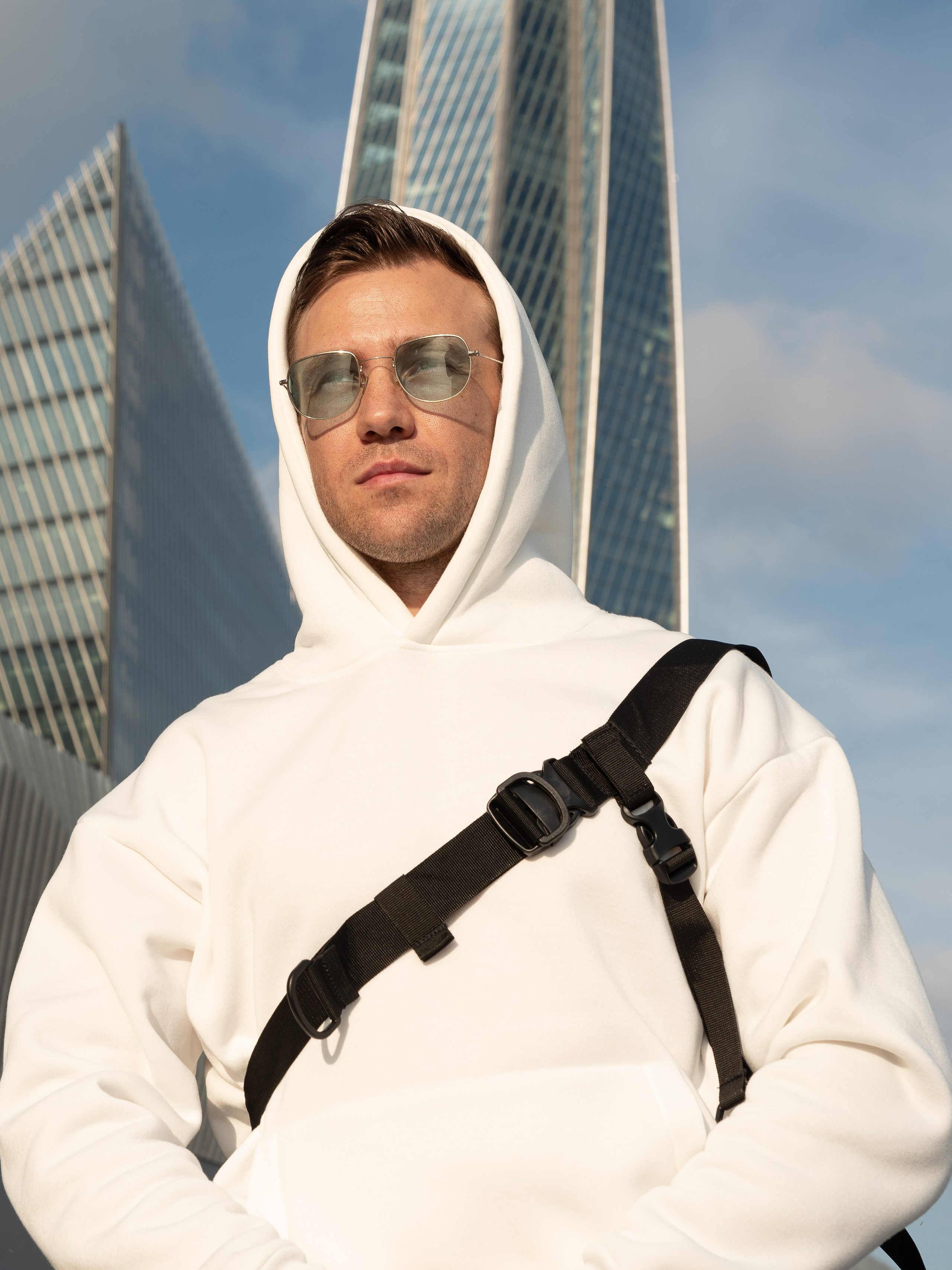 Мужчина в белой кофте с капюшоном и солнечных очках на фоне небоскребов