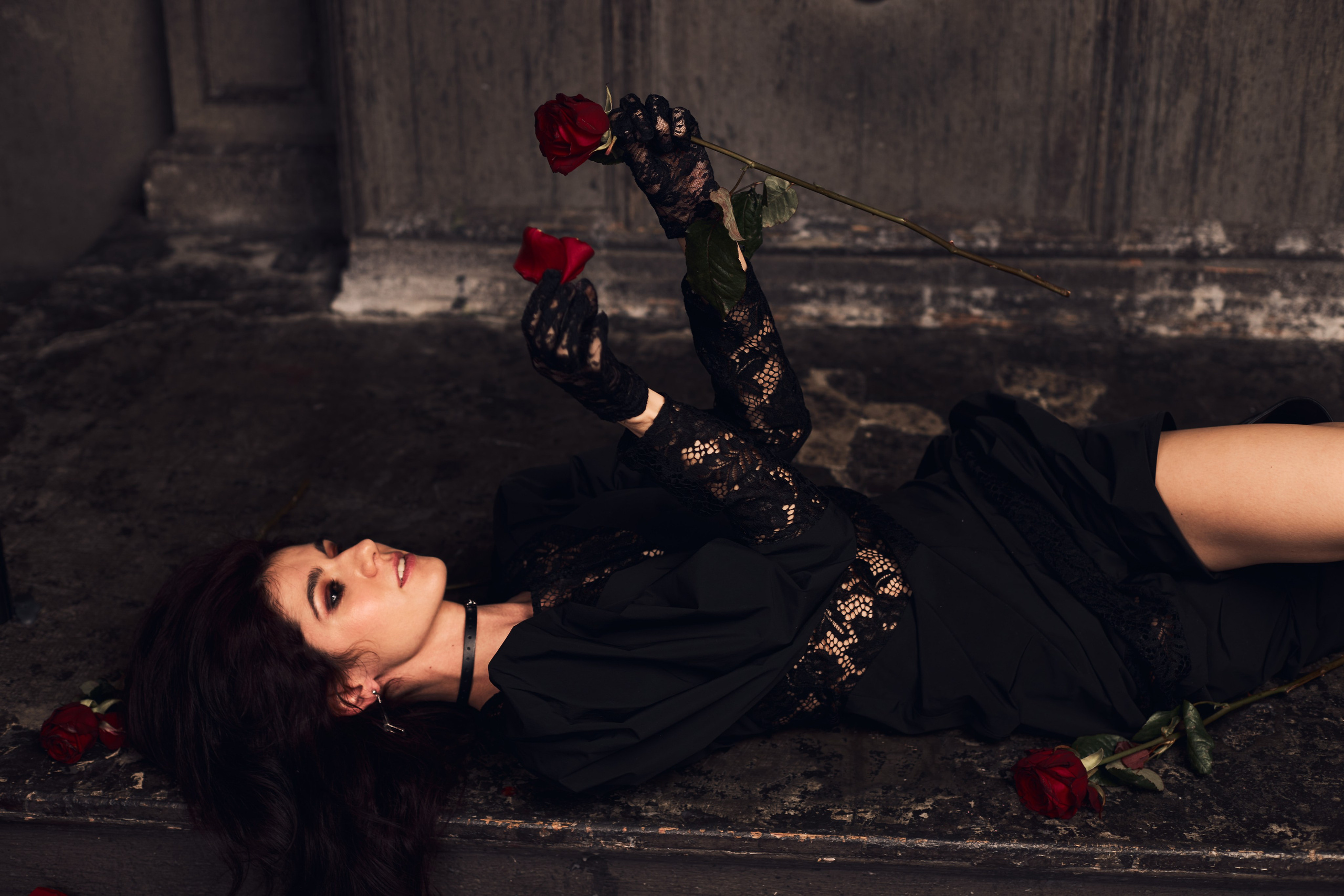 Девушка в черном кружевном платье и в перчатках обрывает лепестки с розы, лёжа на полу