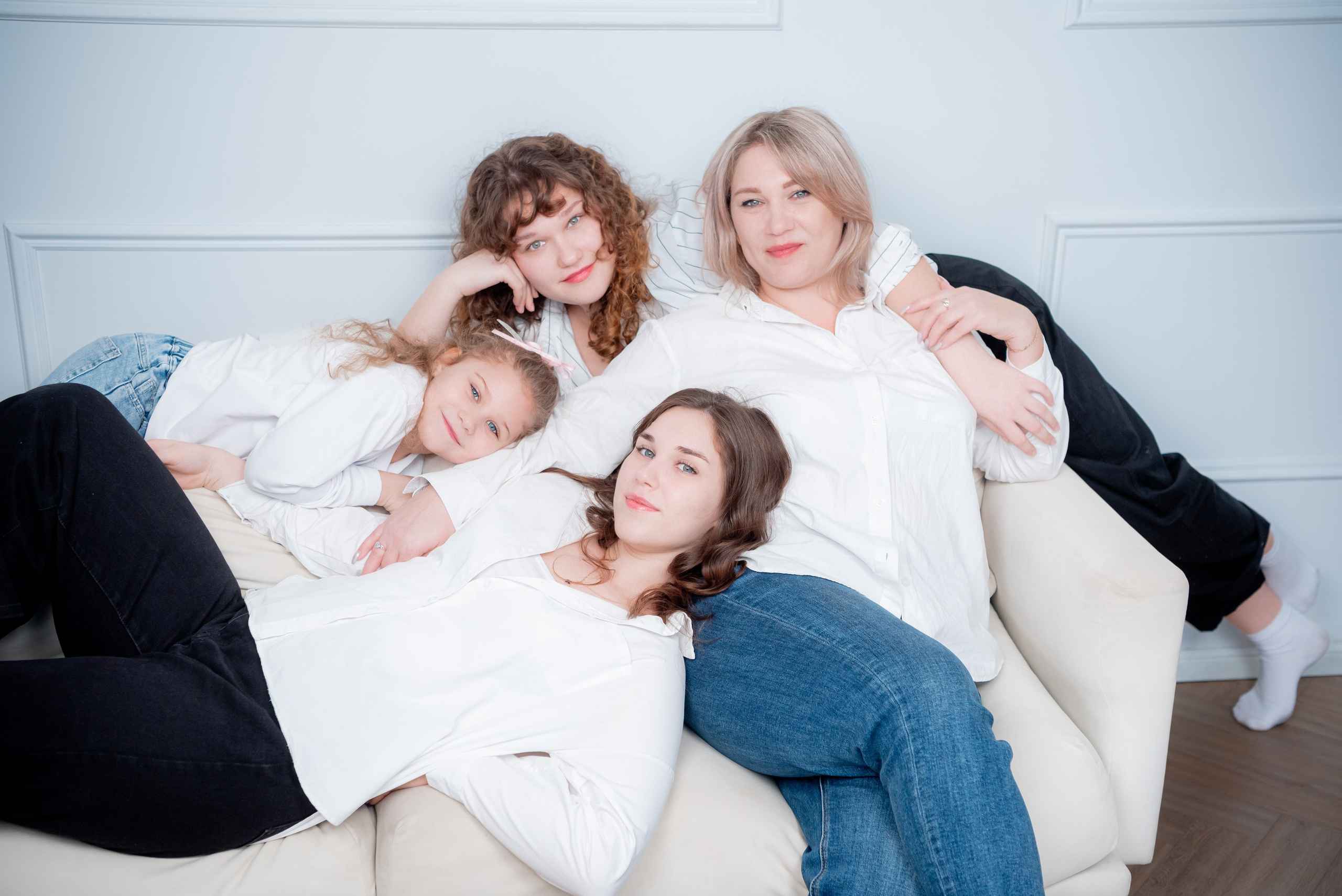 Съемка в студии с большими окнами: мама и три дочки разного возраста сидят на диване, смеются.